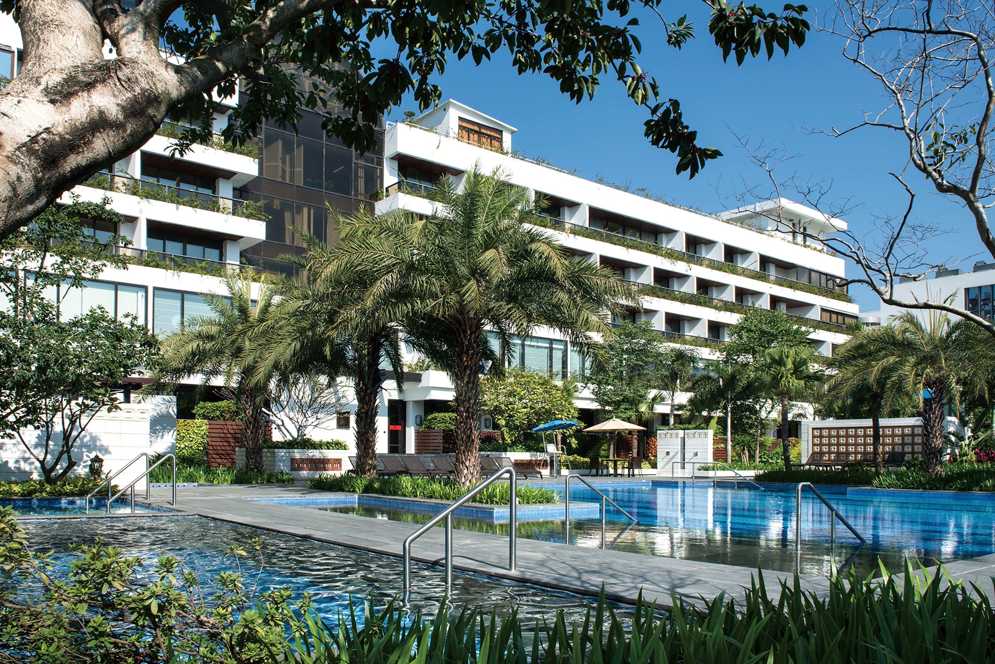 A modern hotel with a swimming pool, palm trees, and a clear blue sky.