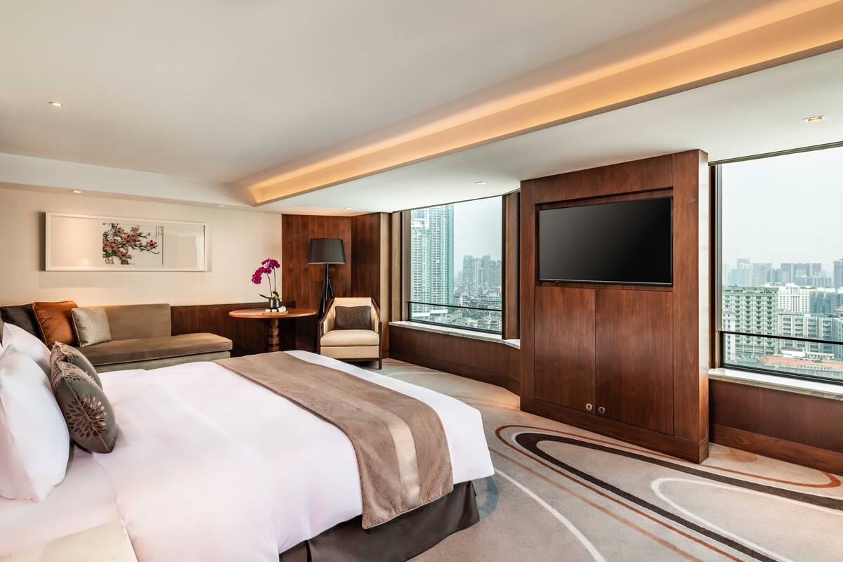 A modern hotel room with a large bed, a TV, a sofa, a small table, and a view of the city.