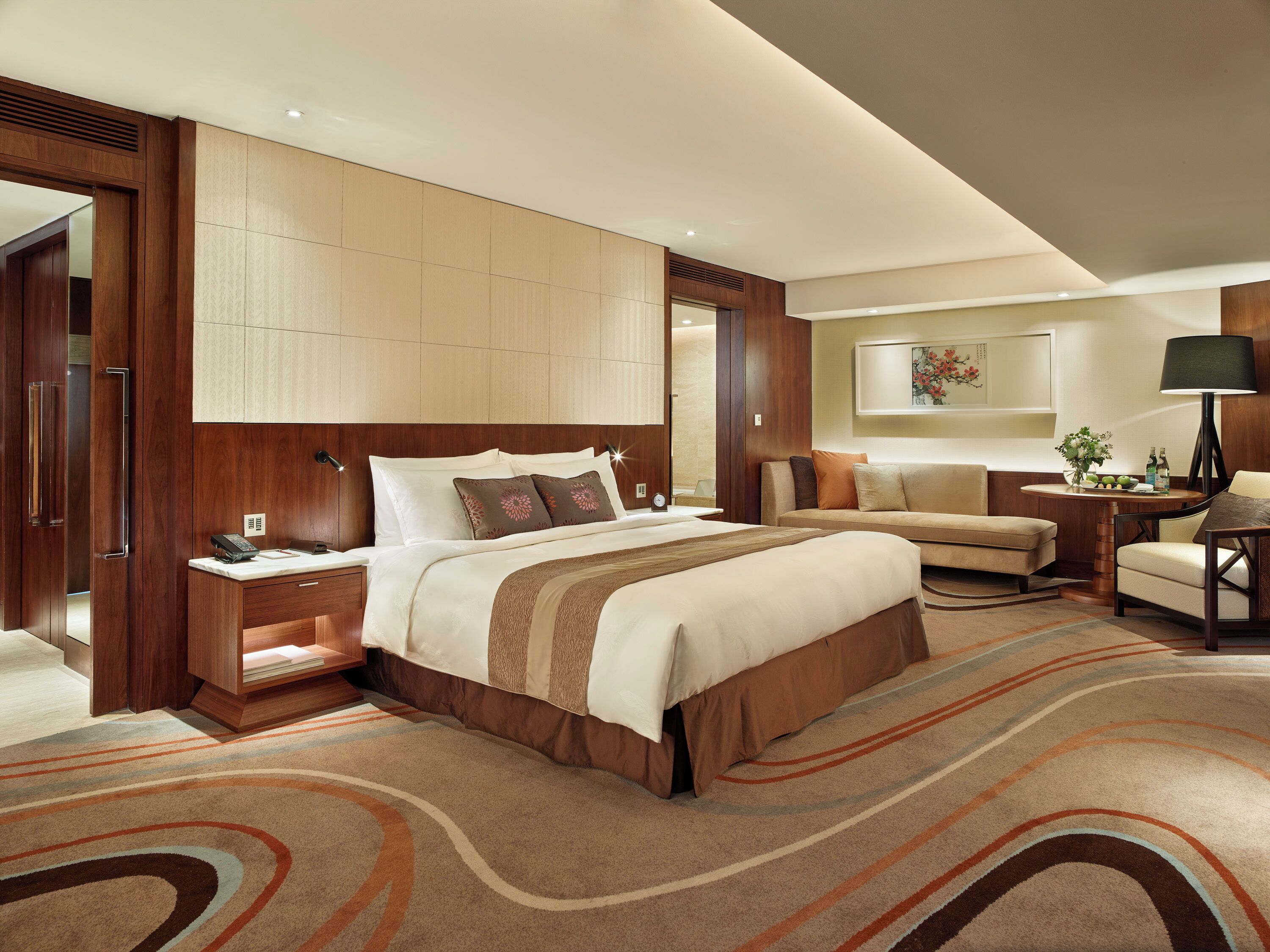 A hotel room with a large bed, a sofa, a desk with a lamp, and a wall-mounted artwork.