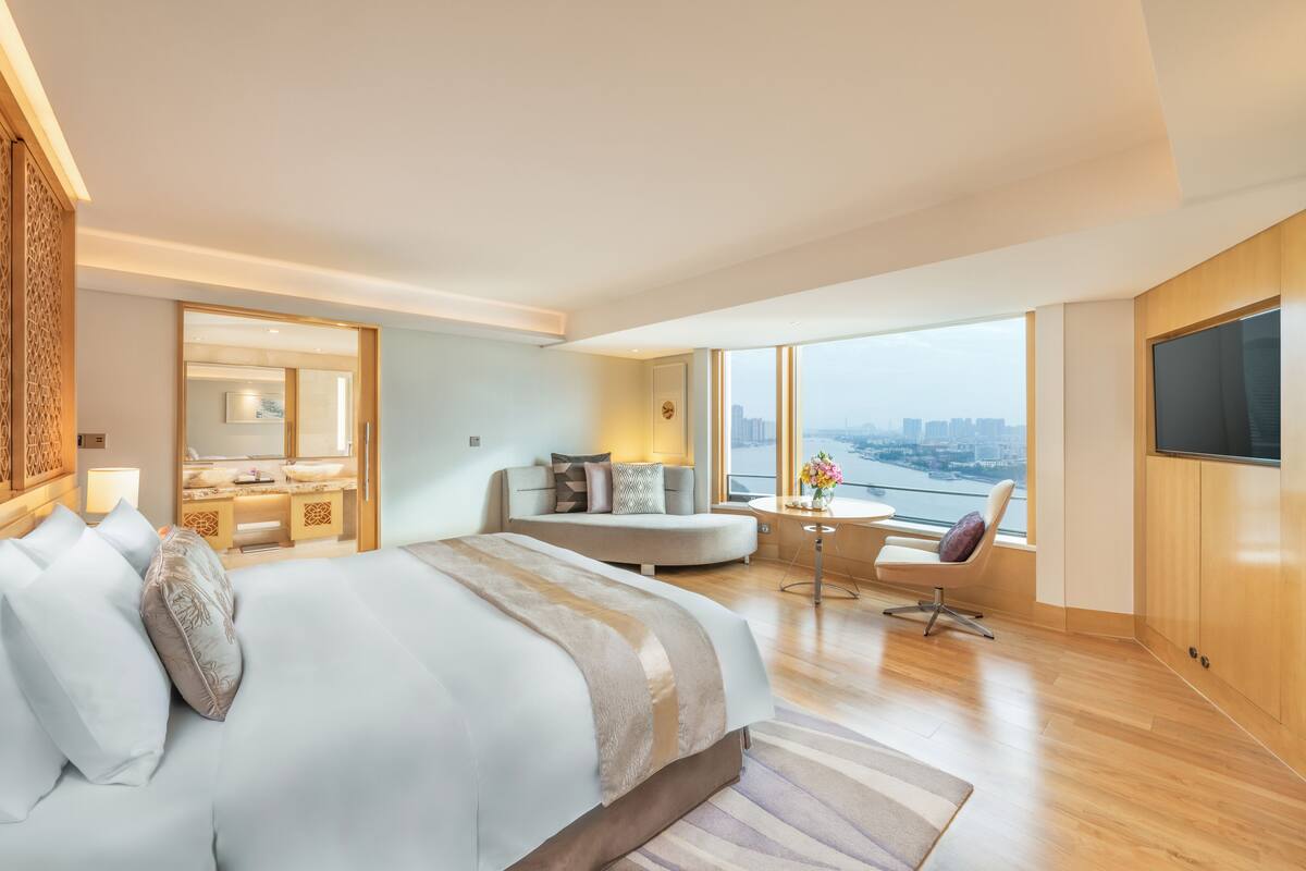 A modern hotel room with a large bed, a sitting area with a view, a desk, and a TV.