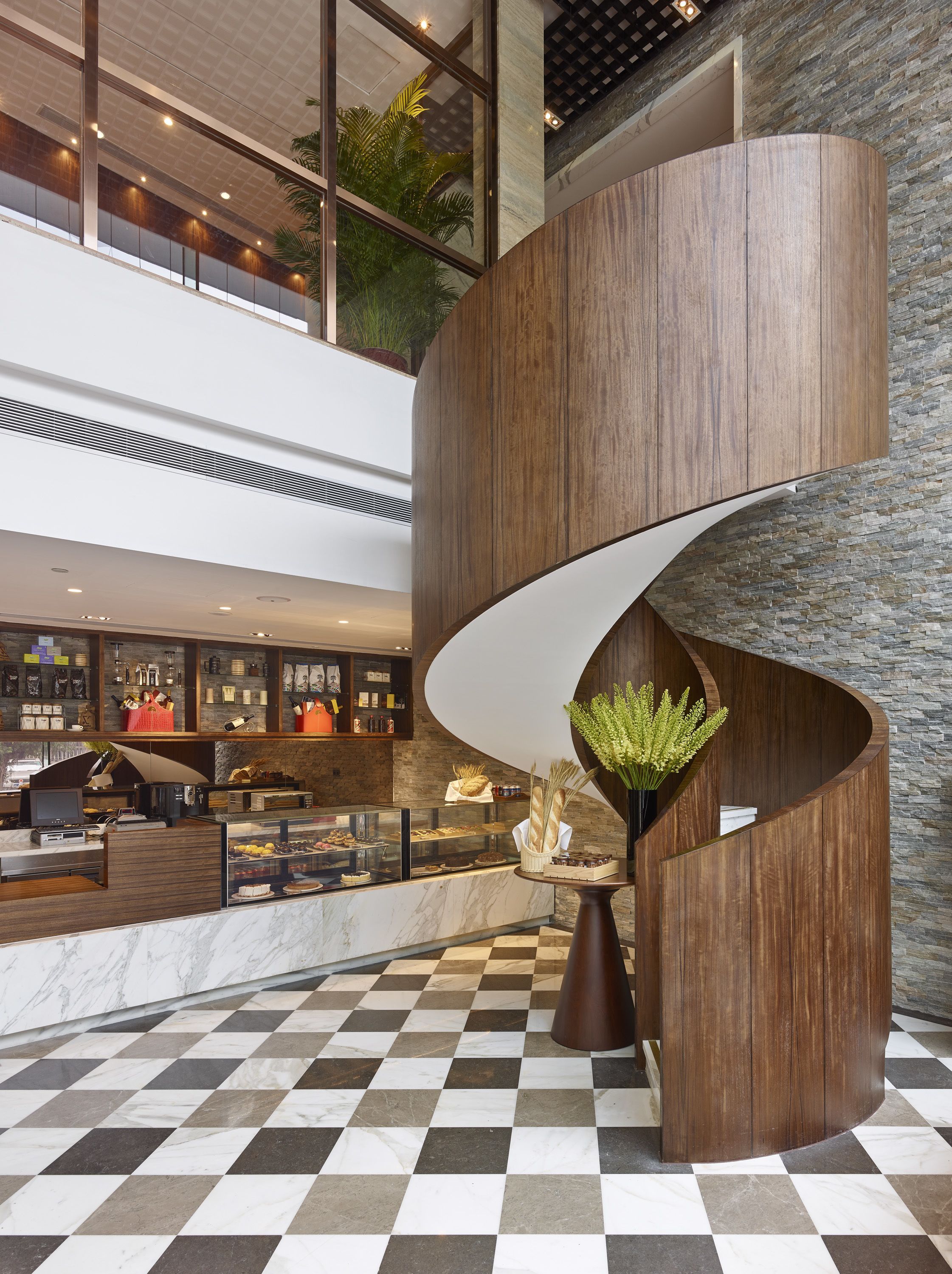 Un hall d’hôtel moderne avec un sol à carreaux, un escalier en bois aux courbes élégantes et un présentoir rempli de pâtisseries et de cafés.