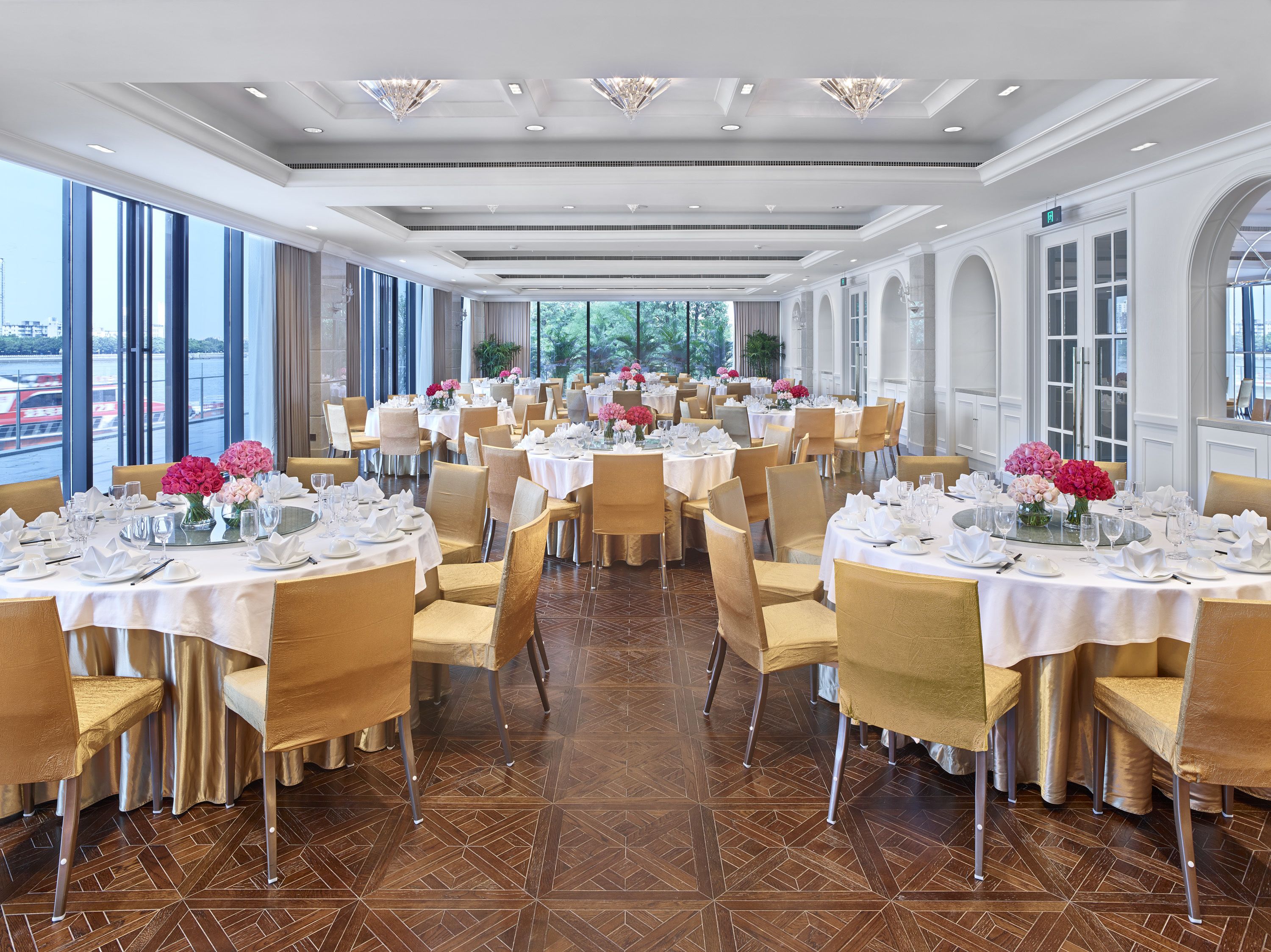 A spacious dining area with multiple tables set for a formal event, large windows offering a view of the outdoors, and a modern ceiling design.