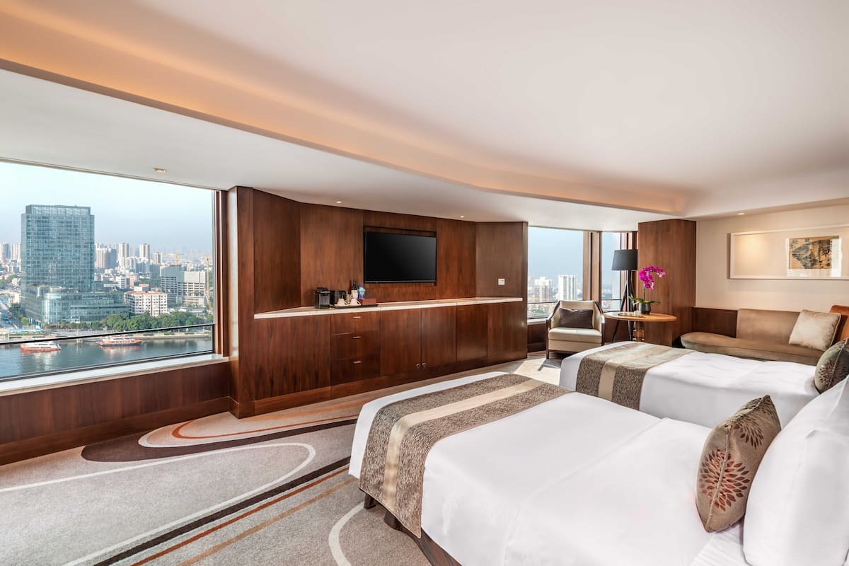 A modern hotel room with a city view, a large flat-screen TV, a sofa, two beds, and a small table with a lamp.