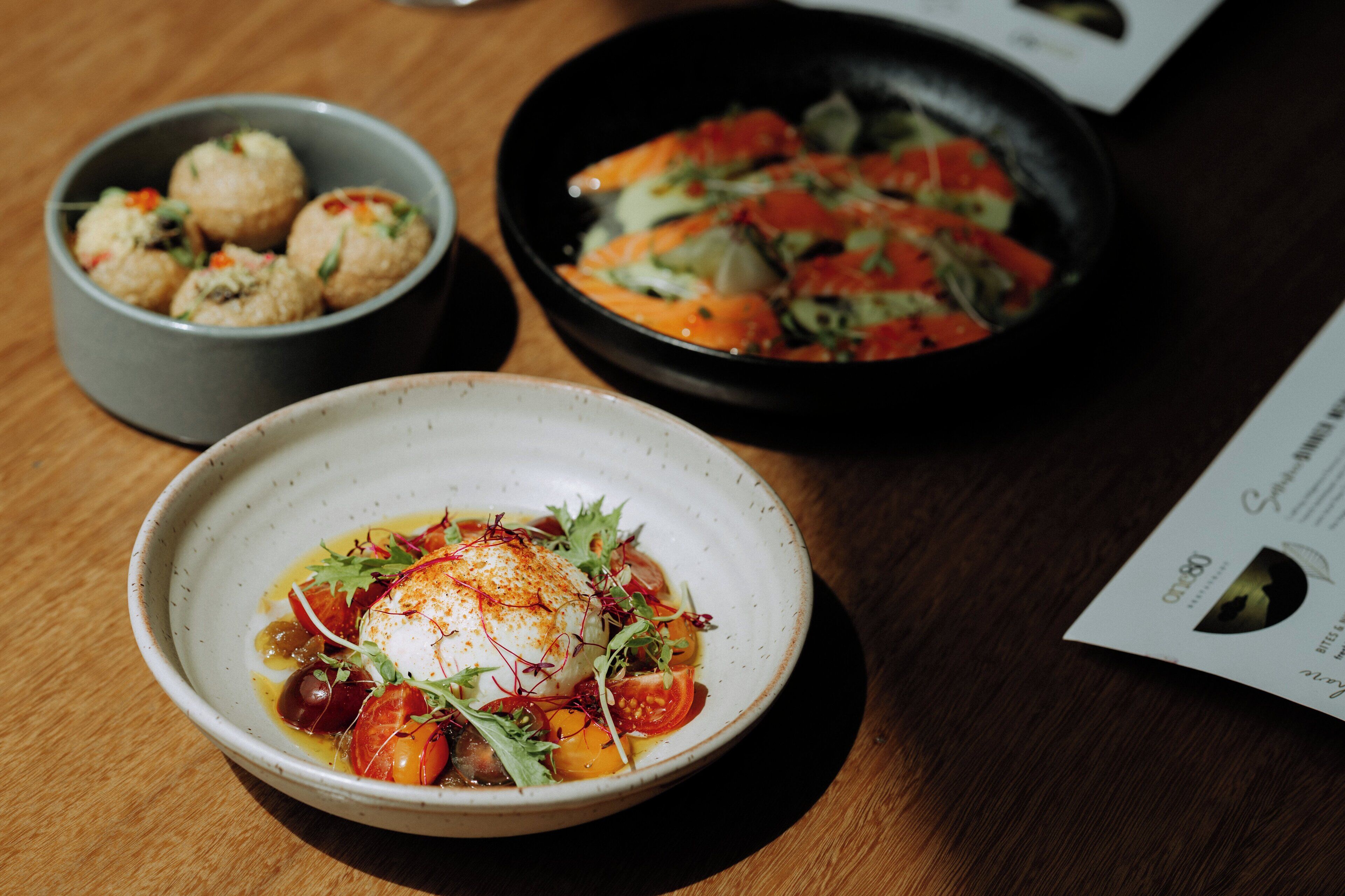 A dining table with three dishes: a bowl of spherical food garnished with herbs, a pan with salmon and vegetables, and a bowl with poached eggs and vegetables.