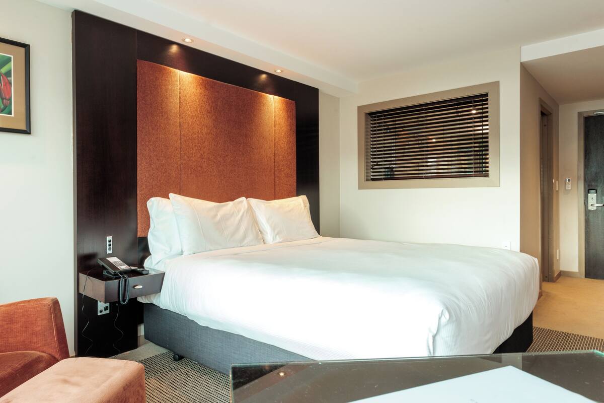 A hotel room with a large bed, a bedside table with a phone, a chair, and a window with blinds.