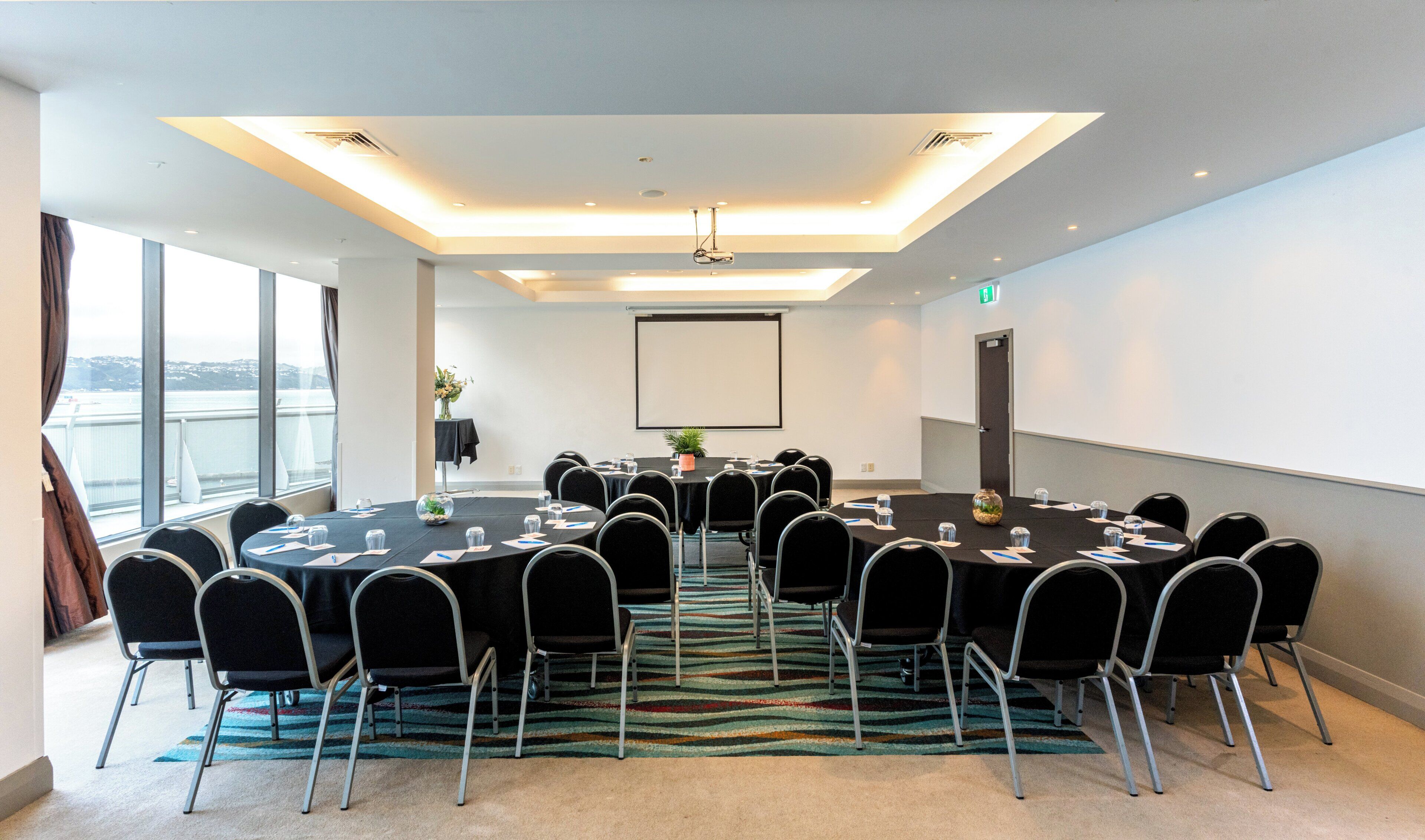 A conference room with a large table, chairs, a projector screen, and a view of the city through large windows.
