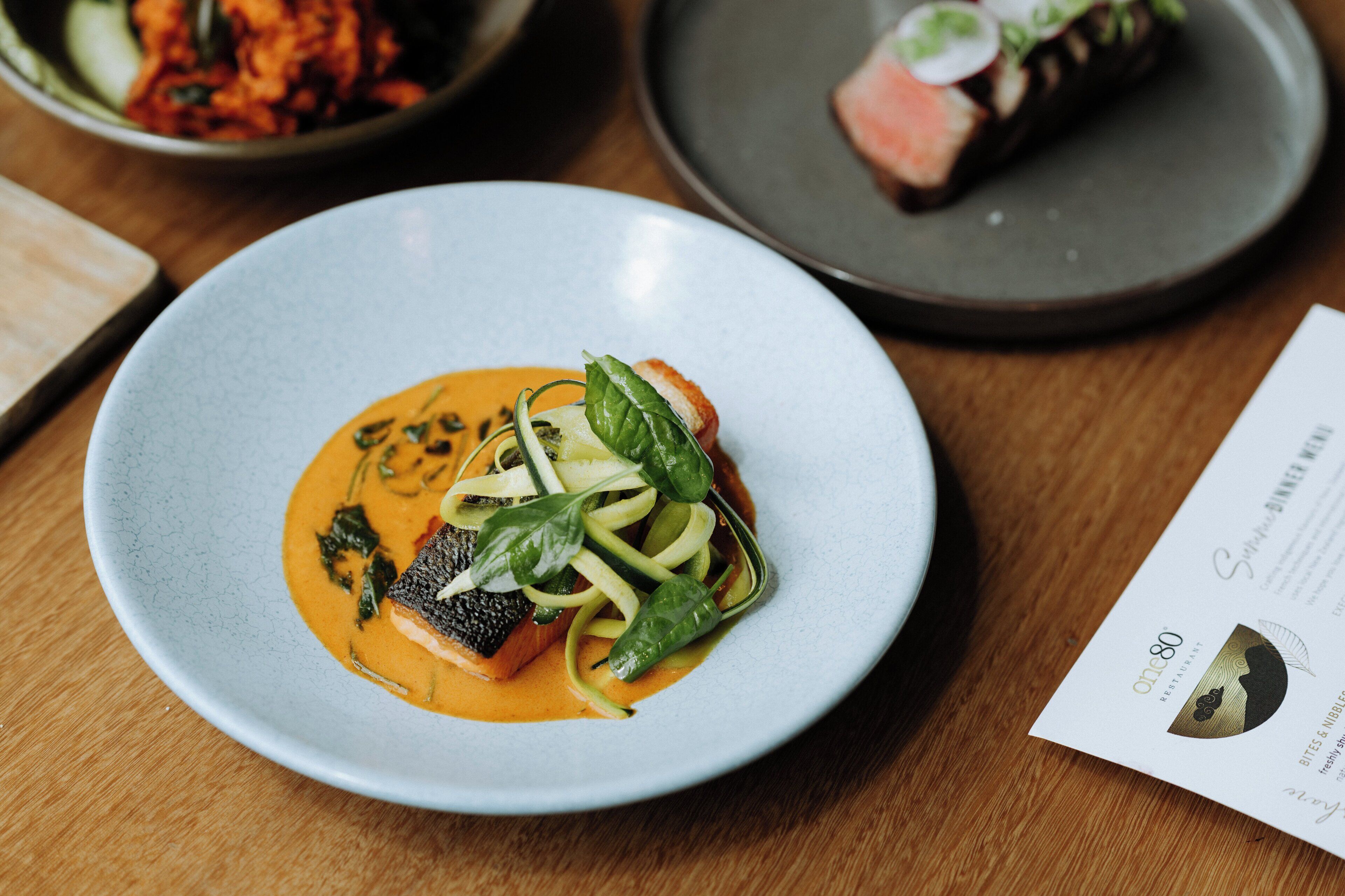 A plate of food with a fish and vegetables, a menu, and a bowl of food in the background.