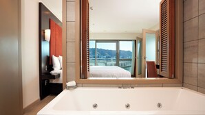 A modern bathroom with a large bathtub, a mirror reflecting a bedroom with a bed and a city view, and a shower area. - Copthorne Hotel Wellington Oriental Bay (Wellington)