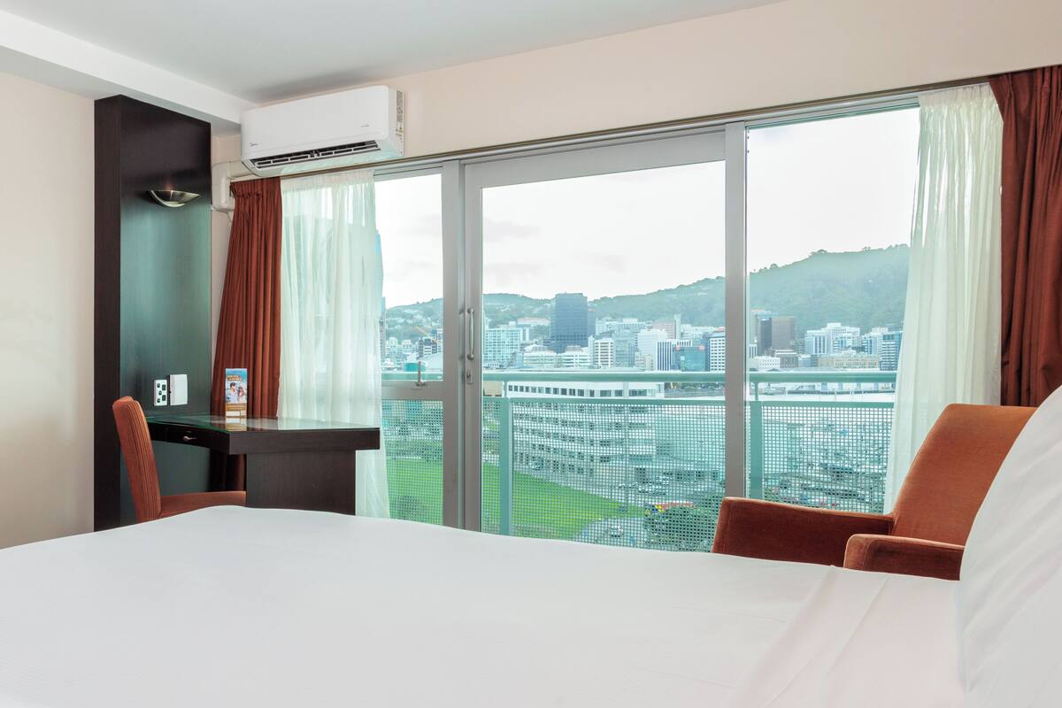 A hotel room with a large bed, a desk, and a chair. There is a window with a view of a cityscape and hills.