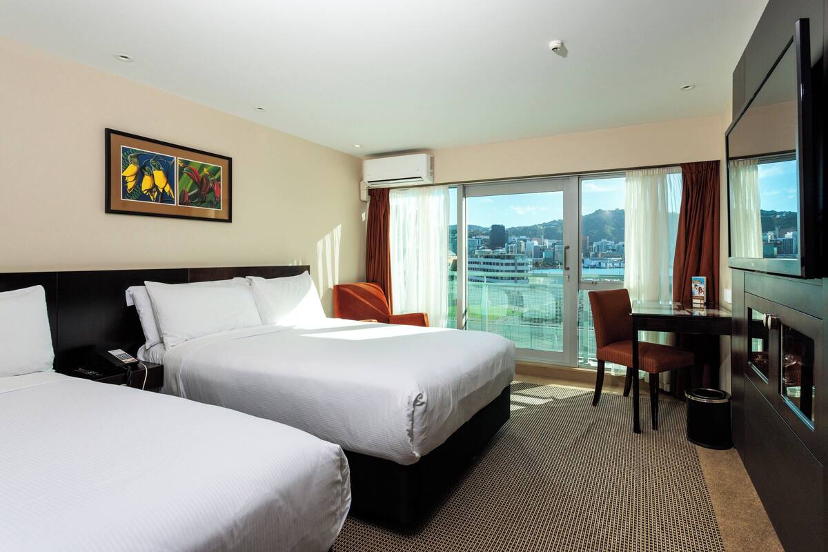 A hotel room with two beds, a desk, a chair, a TV, and a large window with a city view.
