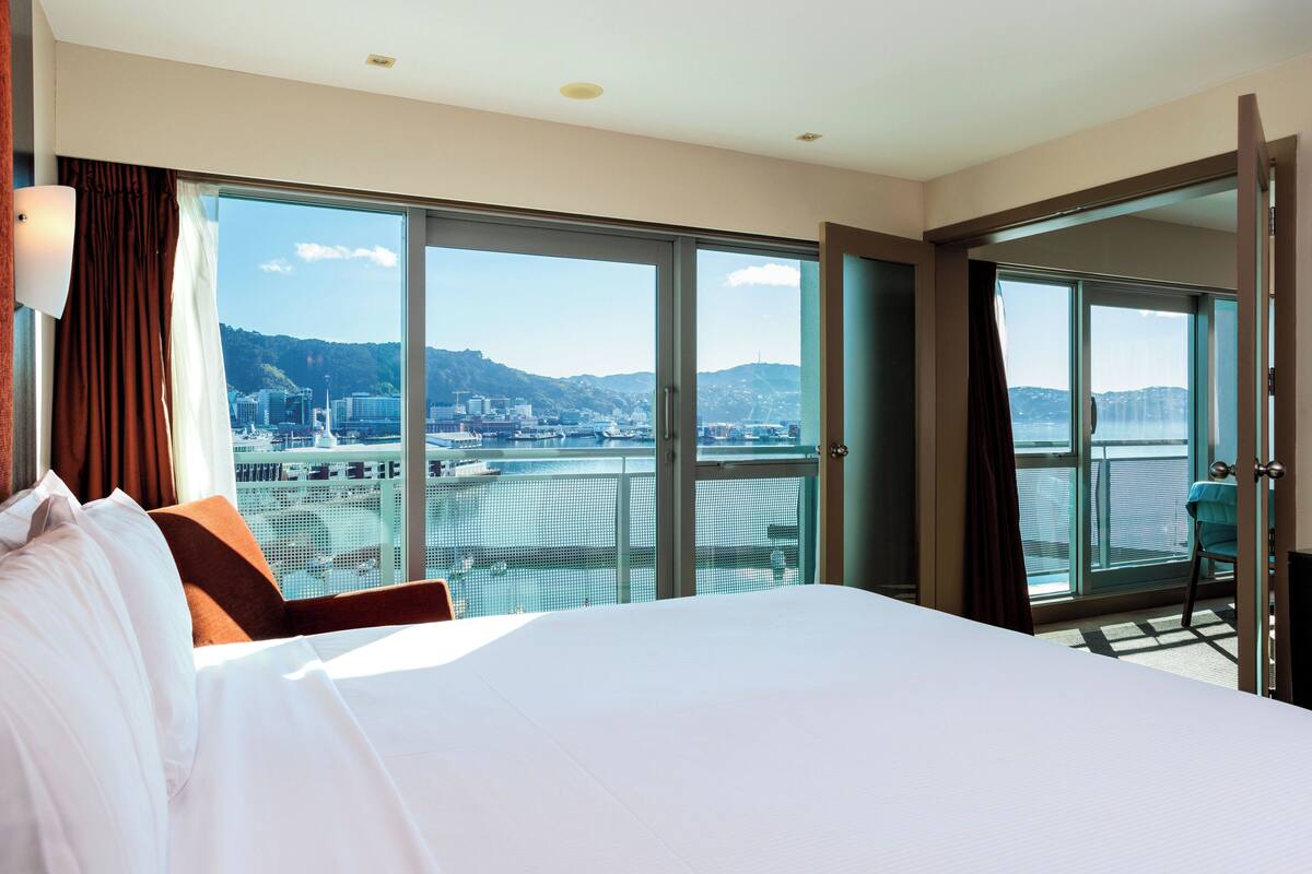 A hotel room with a large bed, a view of a cityscape and water, and a balcony with a chair.