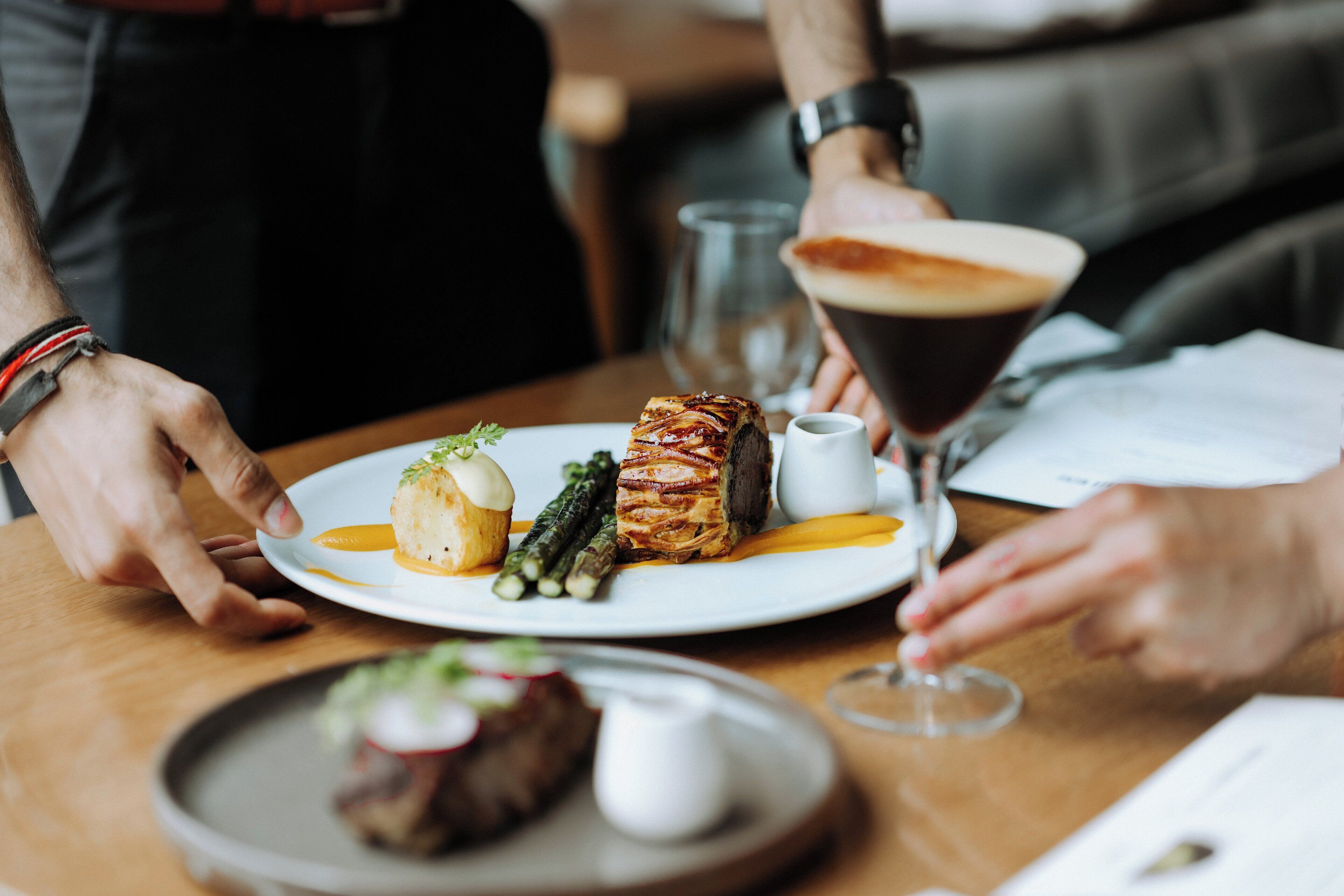 A dining table with a plate of food, a glass of drink, and a person wearing a watch.
