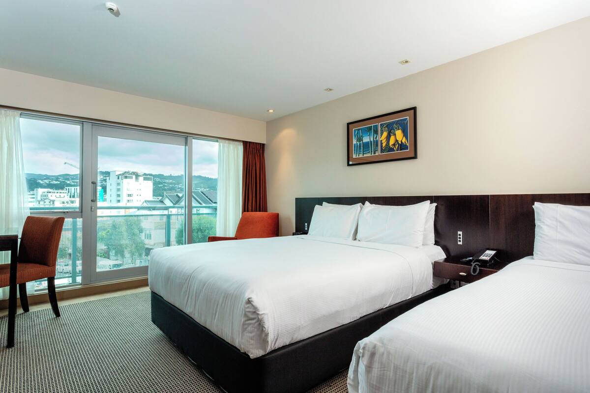 A hotel room with two beds, a desk, and a large window with a view of buildings and greenery.