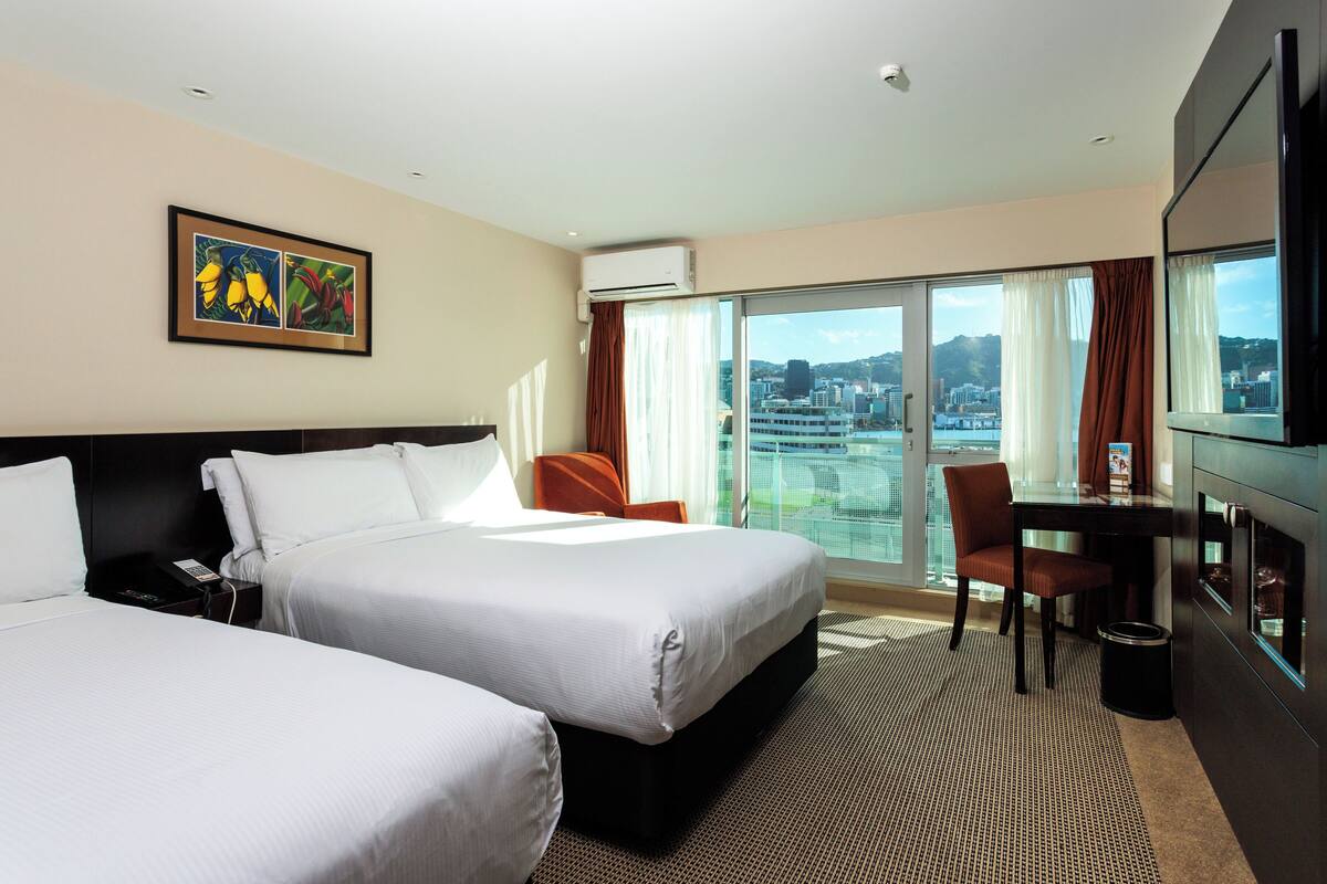 A hotel room with two beds, a desk, a chair, a TV, and a large window with a city view.