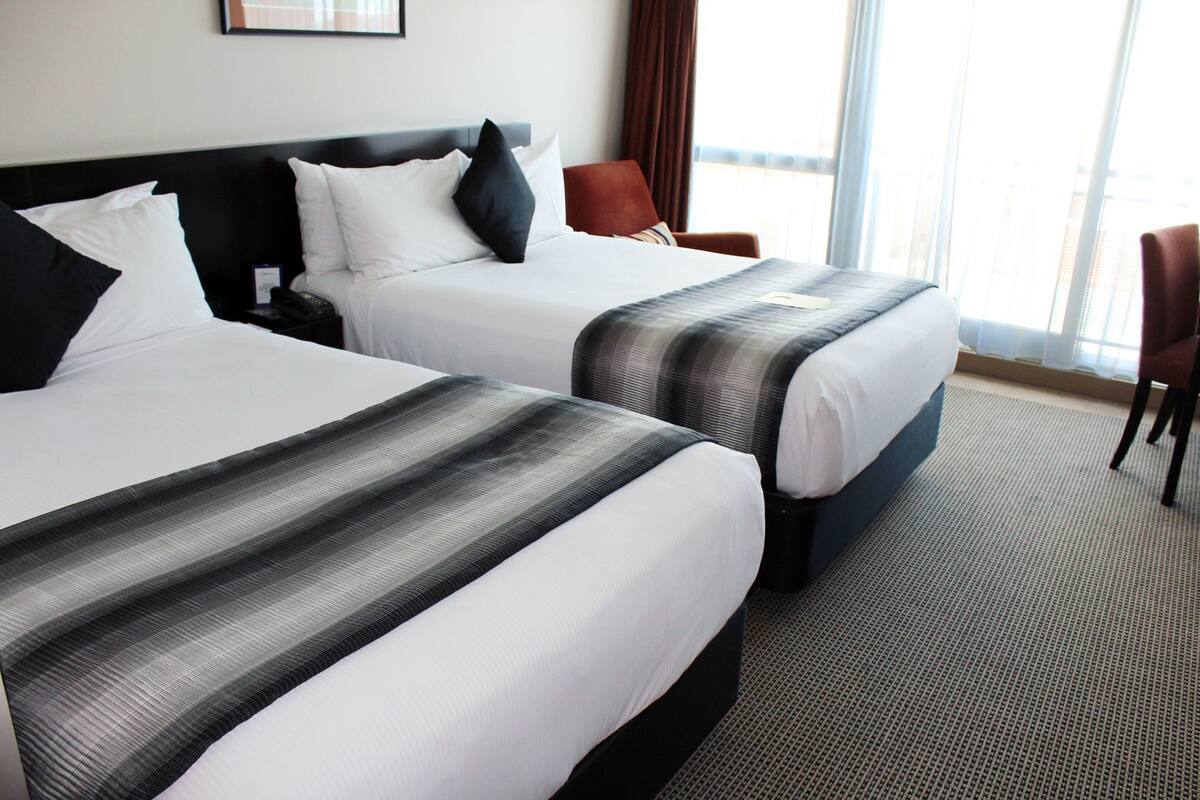 A hotel room with two beds, a nightstand, a chair, and a large window.