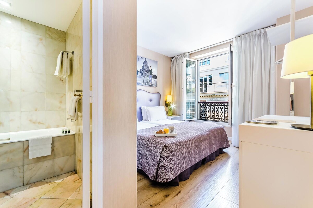 Une chambre d’hôtel moderne dotée d’un grand lit, d’une salle de bain avec baignoire et d’un balcon offrant une vue sur la ville.