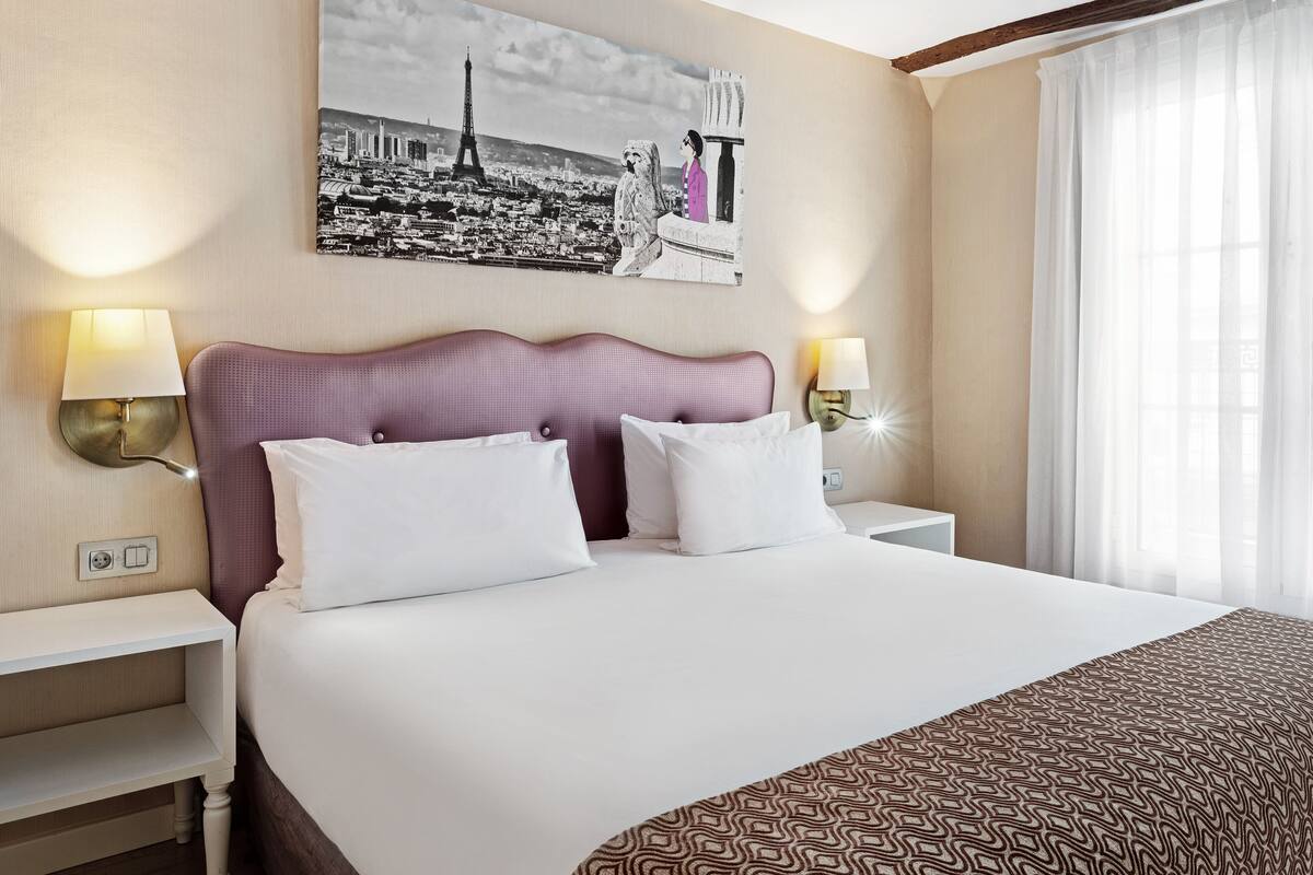 A hotel room with a large bed, two bedside tables, a wall-mounted lamp, and a cityscape painting.