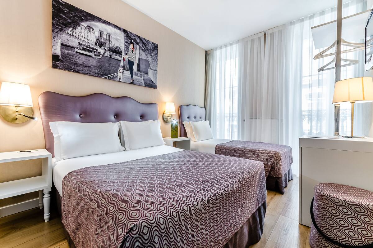 A hotel room with two beds, a large window with sheer curtains, a wall-mounted artwork, and bedside lamps.