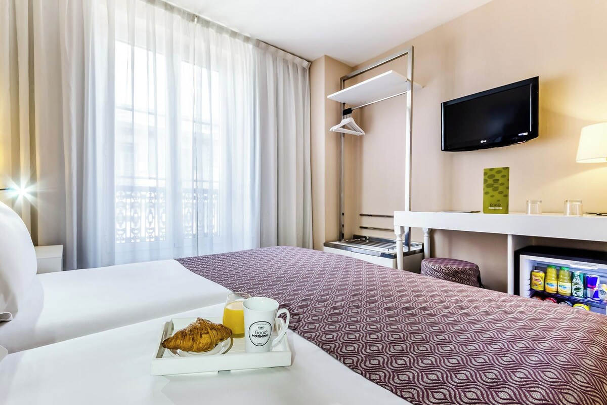 A hotel room with a bed, a TV, a minibar, and a breakfast tray with croissant and coffee.