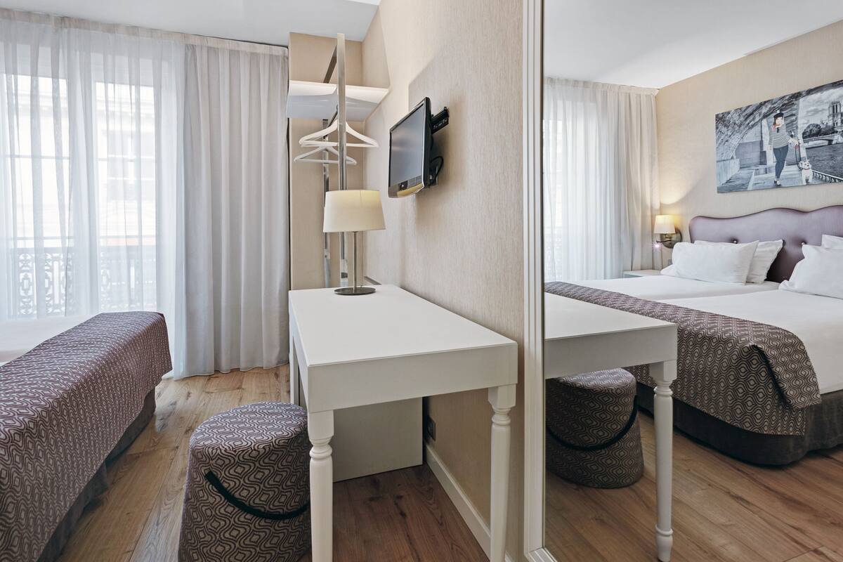 A hotel room with a bed, a desk, a chair, a TV, and a painting on the wall.