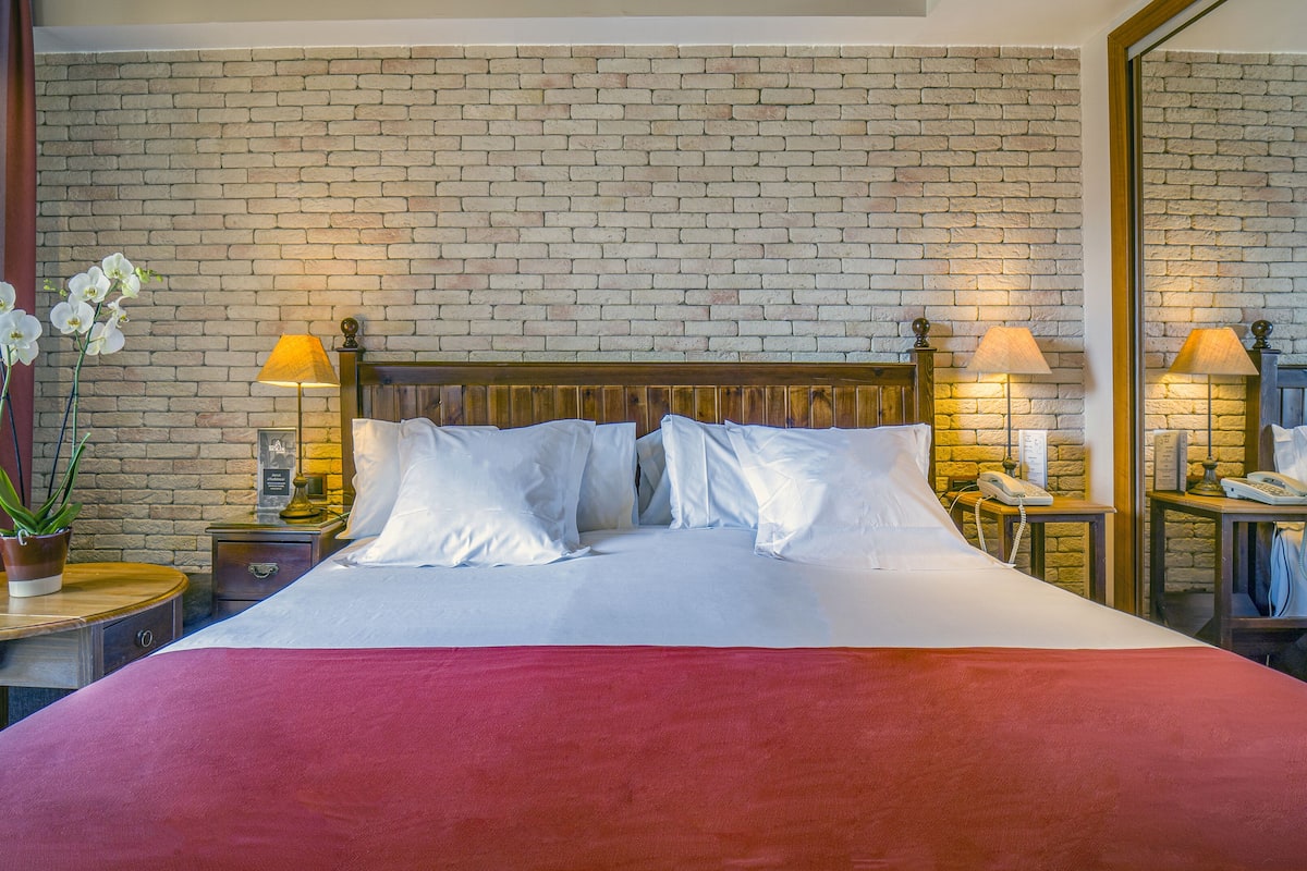 Quarto de hotel com parede de tijolos, estrutura de cama de madeira, roupa de cama branca e duas mesas de cabeceira com abajures e telefone.