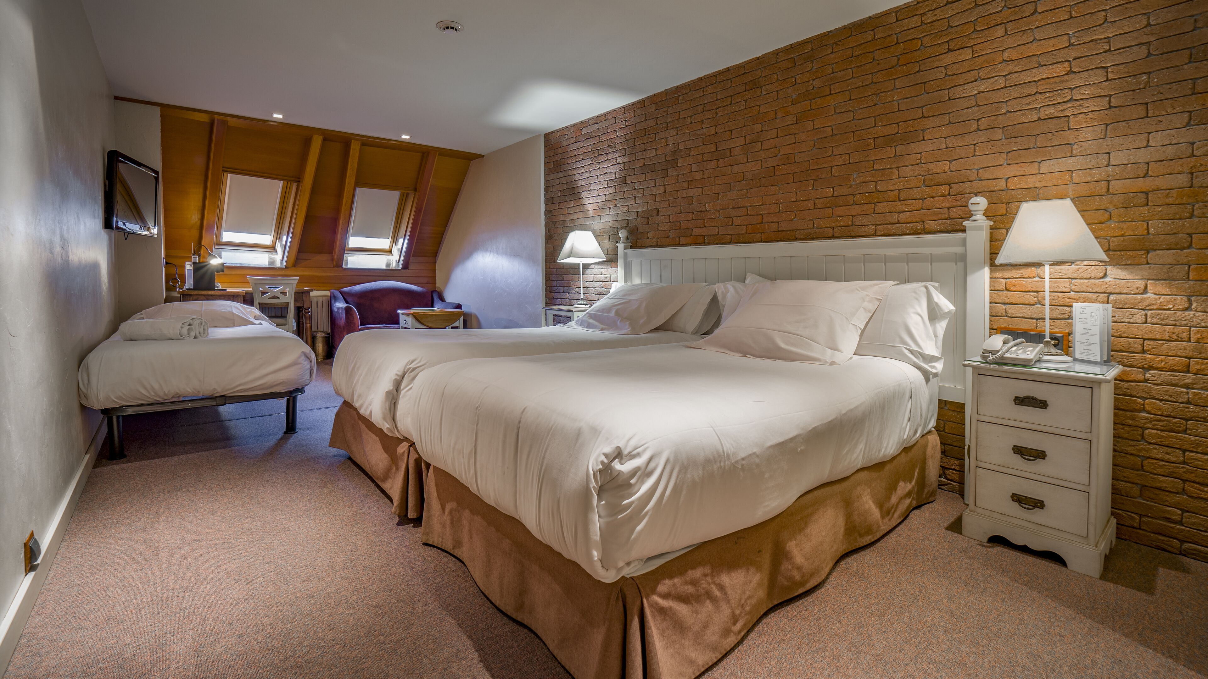 A hotel room with two beds, a brick wall, a wooden ceiling, and a small desk with a lamp.