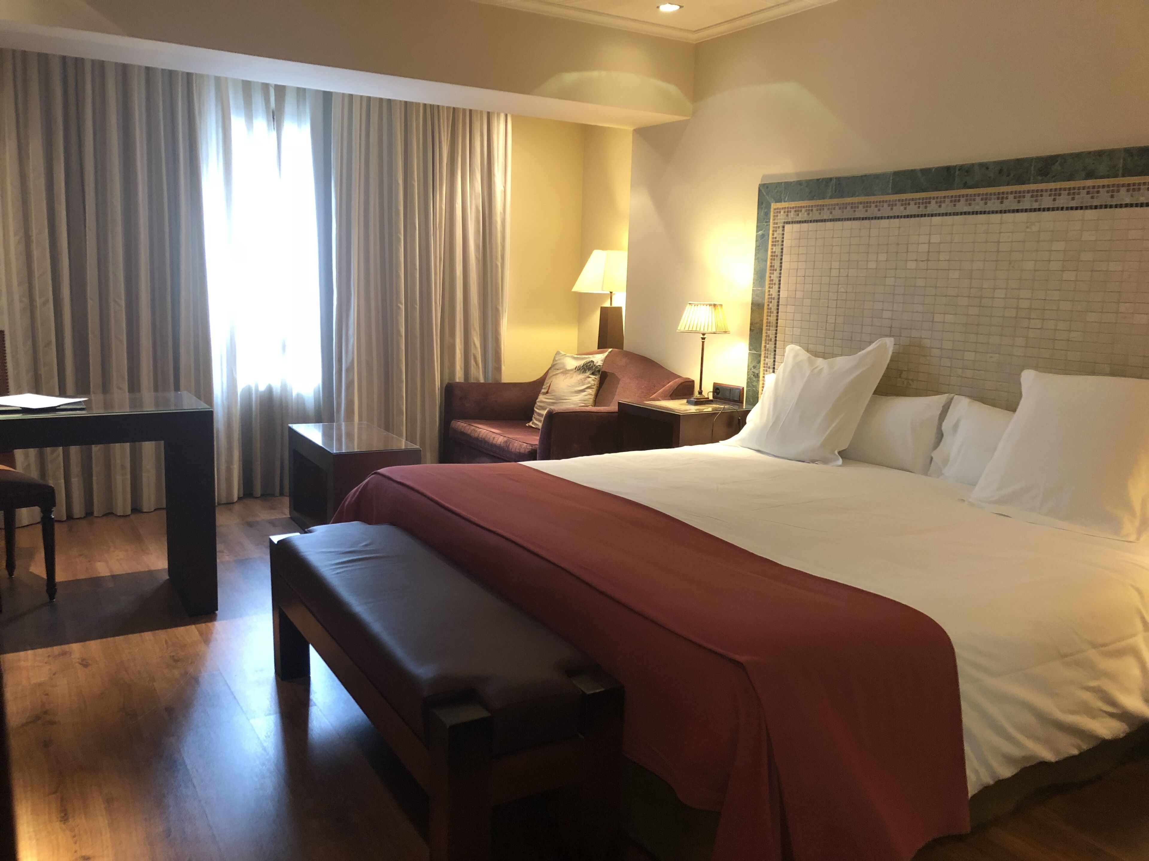 Habitación de hotel con una cama grande, un escritorio, una silla, un banco y una ventana con cortinas.