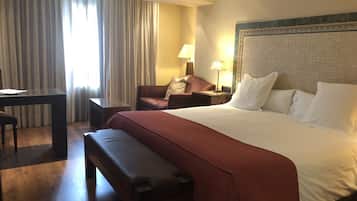 Habitación de hotel con una cama grande, un escritorio, una silla, un banco y una ventana con cortinas.