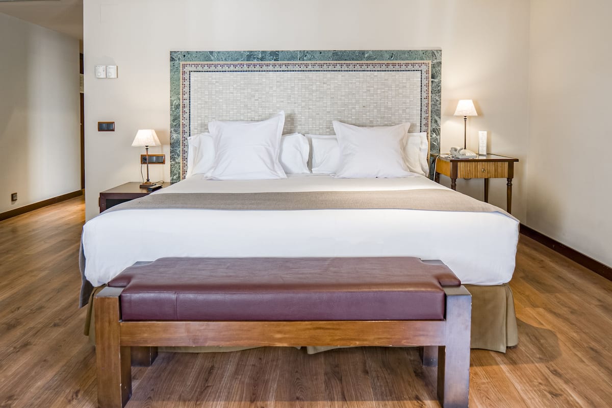 Quarto de hotel com uma cama grande, duas mesas de cabeceira com luminárias, um banco de madeira e uma cadeira.