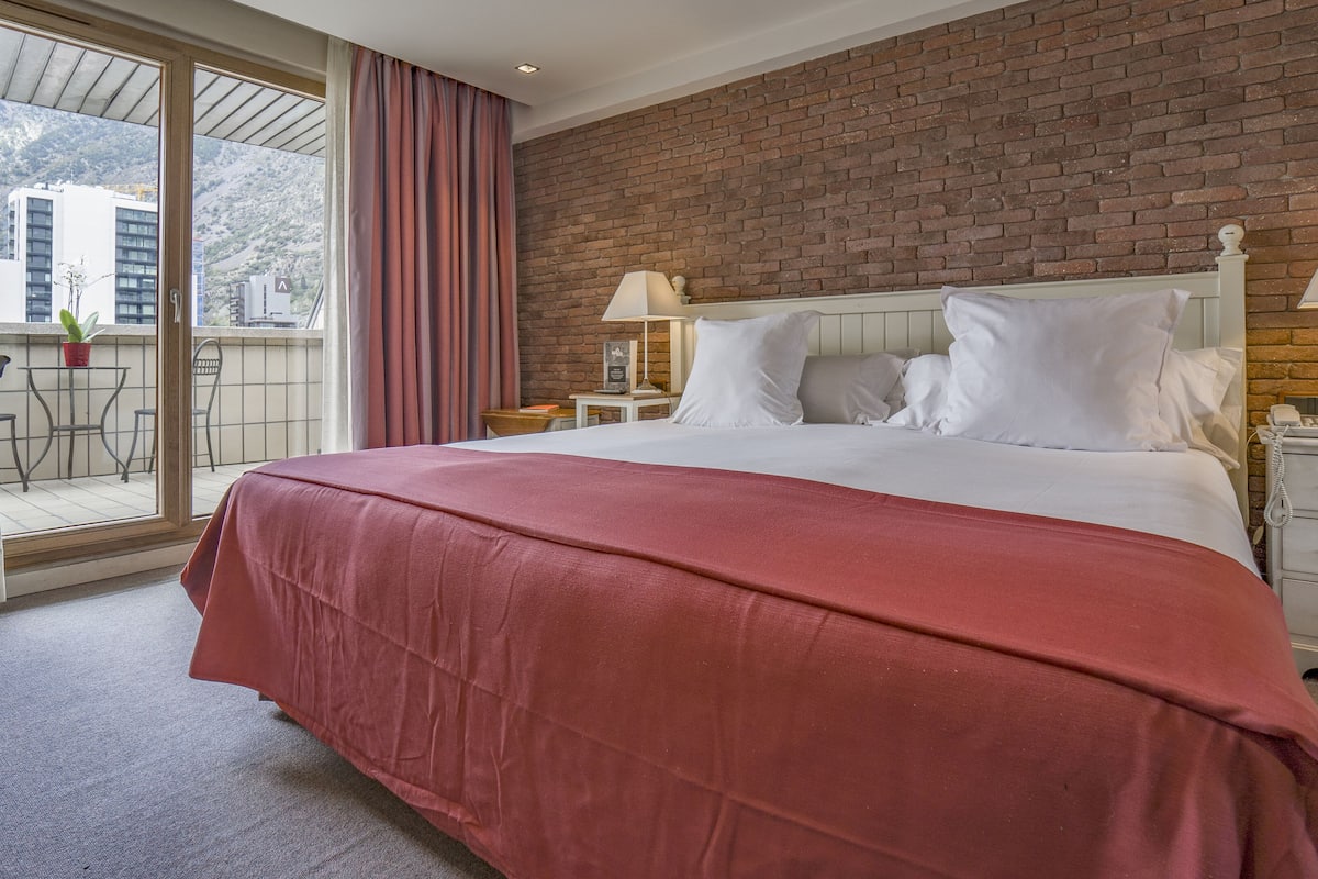 Quarto de hotel com uma cama grande, duas mesinhas de cabeceira, uma parede de tijolos e uma varanda com vista para edifícios e montanhas.