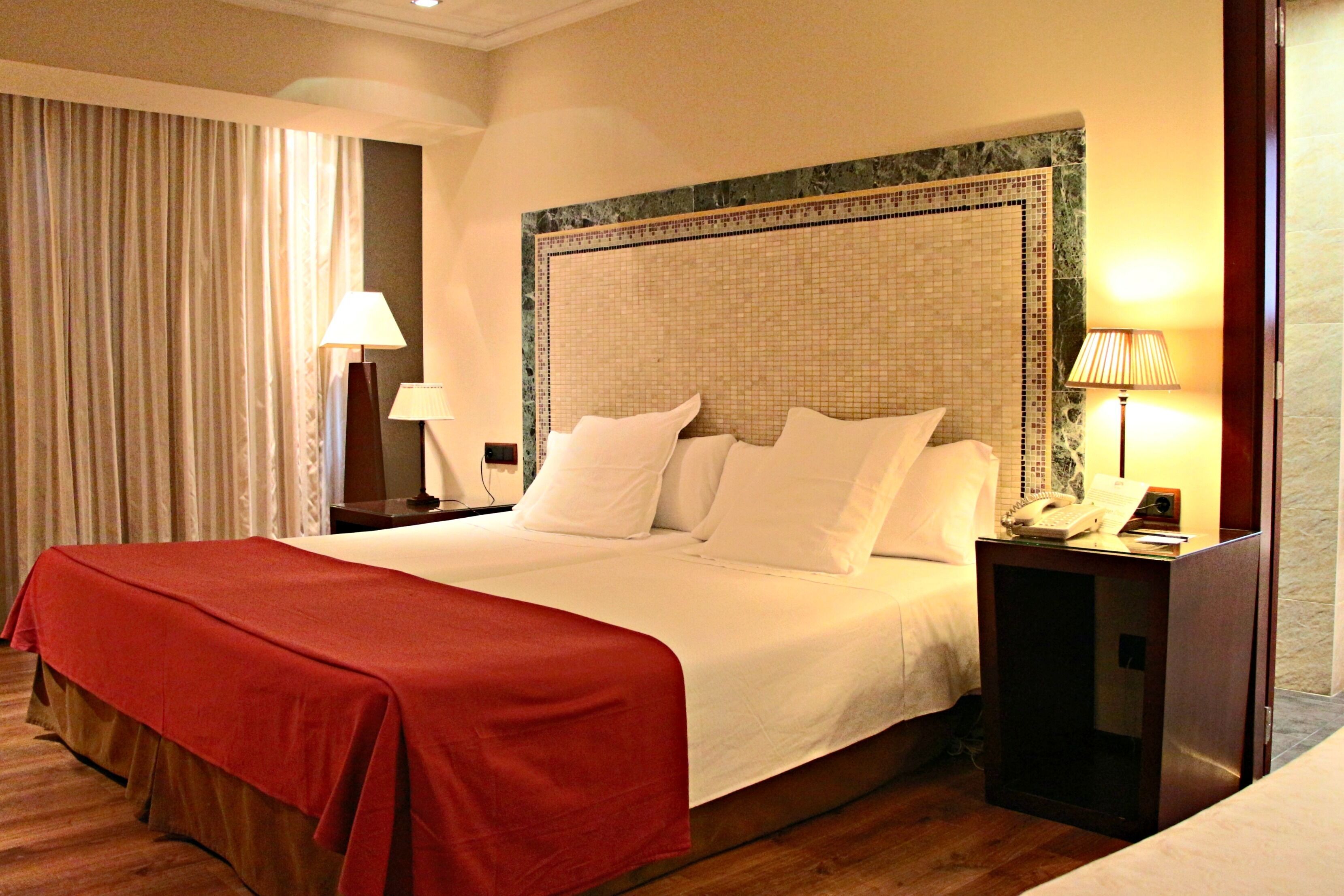 A hotel room with a large bed, a nightstand with a lamp, a telephone, and a red bedspread.