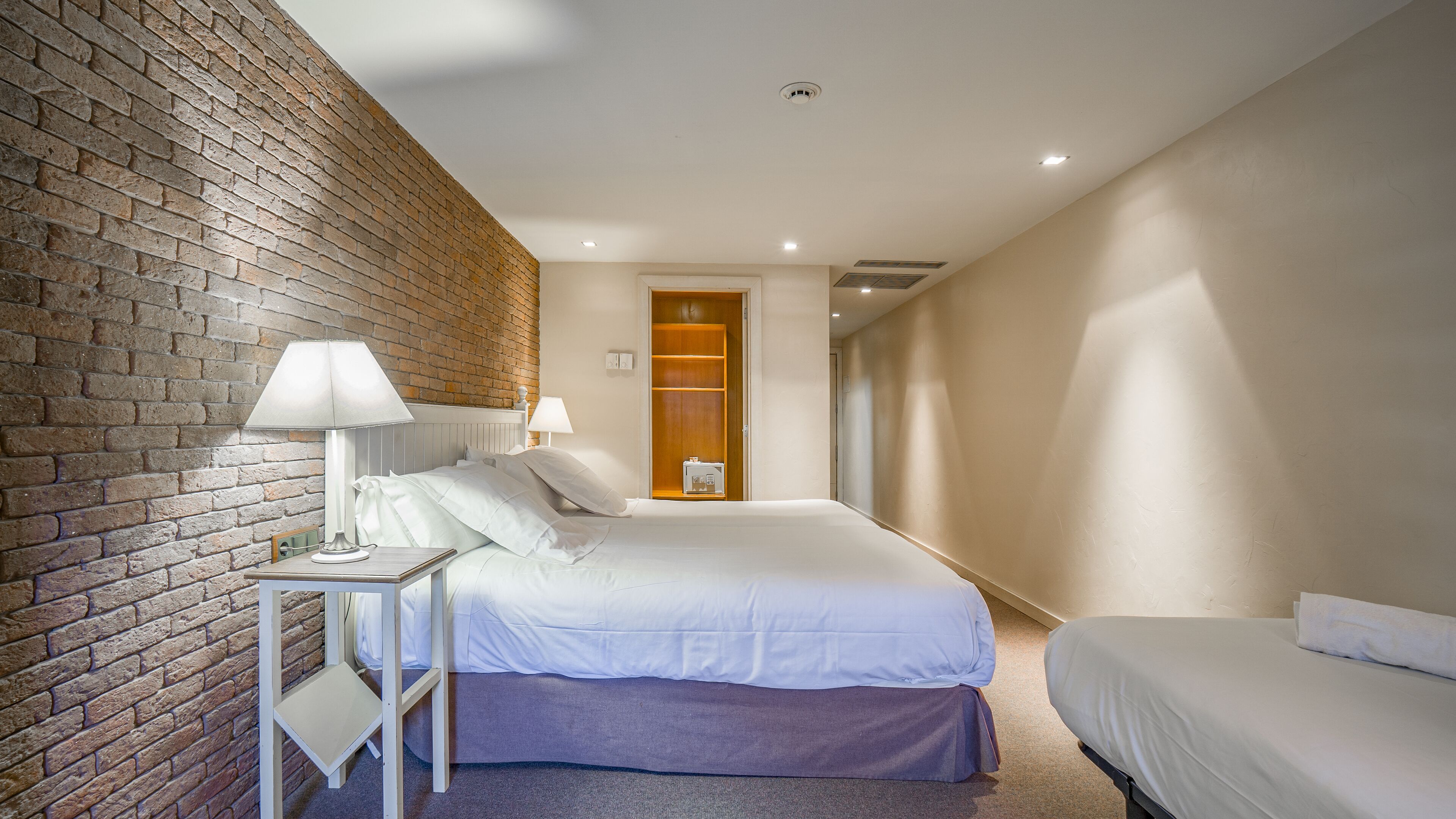 A hotel room with a brick wall, a bed with white bedding, a nightstand with a lamp, and a small table with a chair.