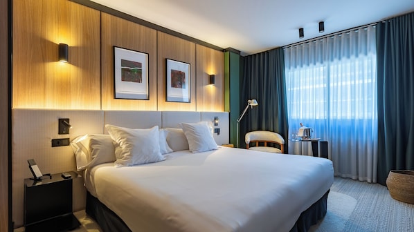 A hotel room with a large bed, a bedside table with a lamp, a chair, a small table with a bottle and glasses, and two framed pictures on the wall.