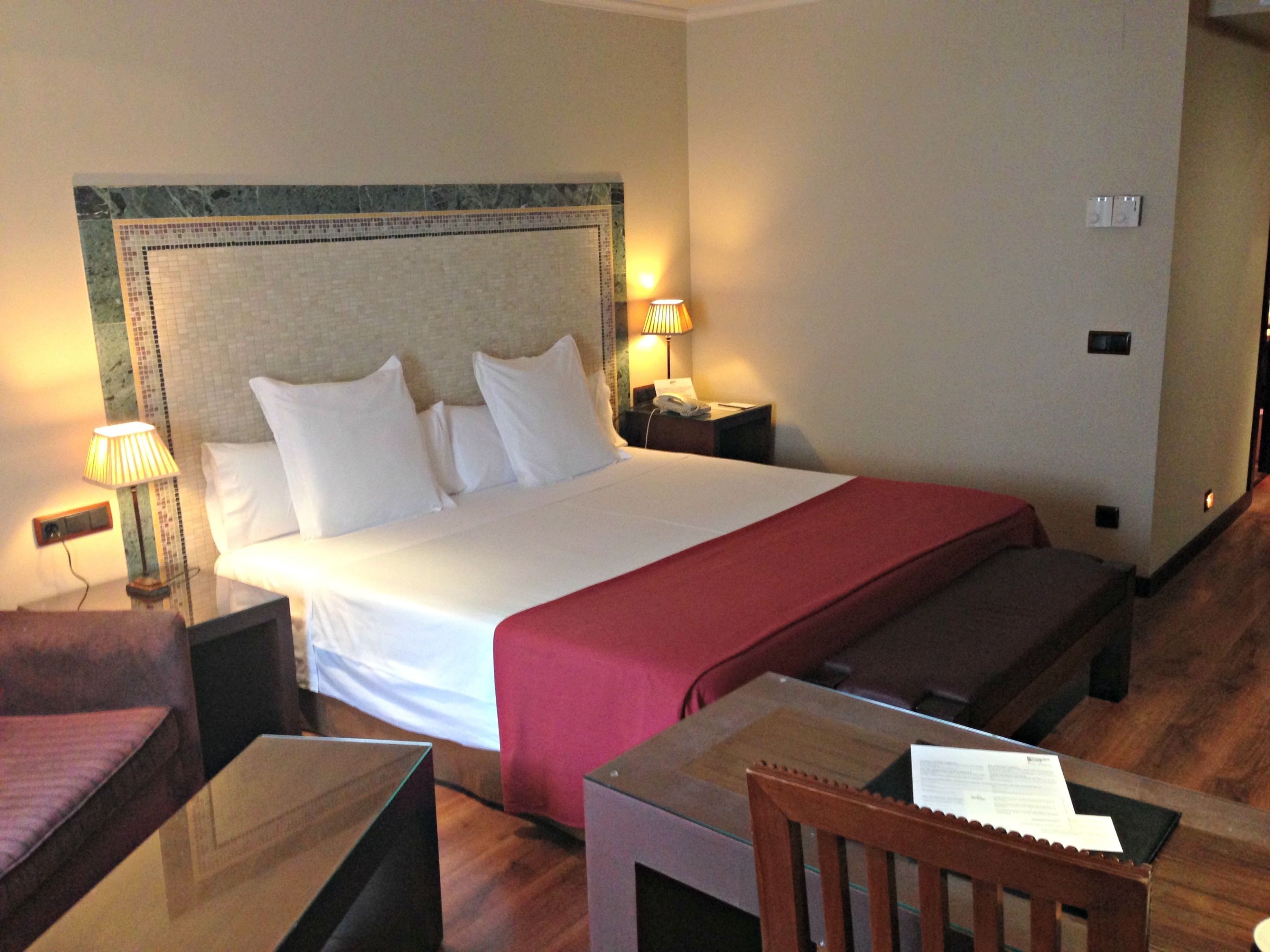 Una habitación de hotel con una cama grande, una mesita de noche, una silla, una pequeña mesa y una lámpara.