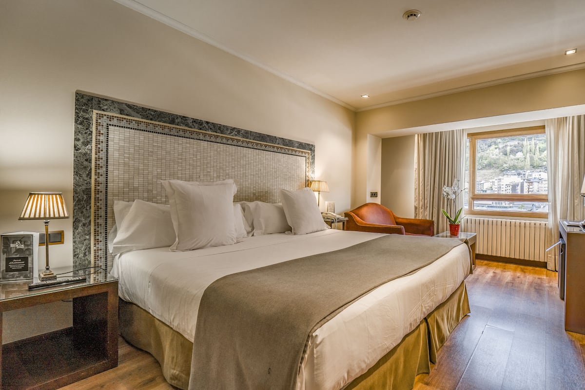 Quarto de hotel com uma cama grande, uma escrivaninha com luminária, uma cadeira, uma televisão e uma janela com vista.