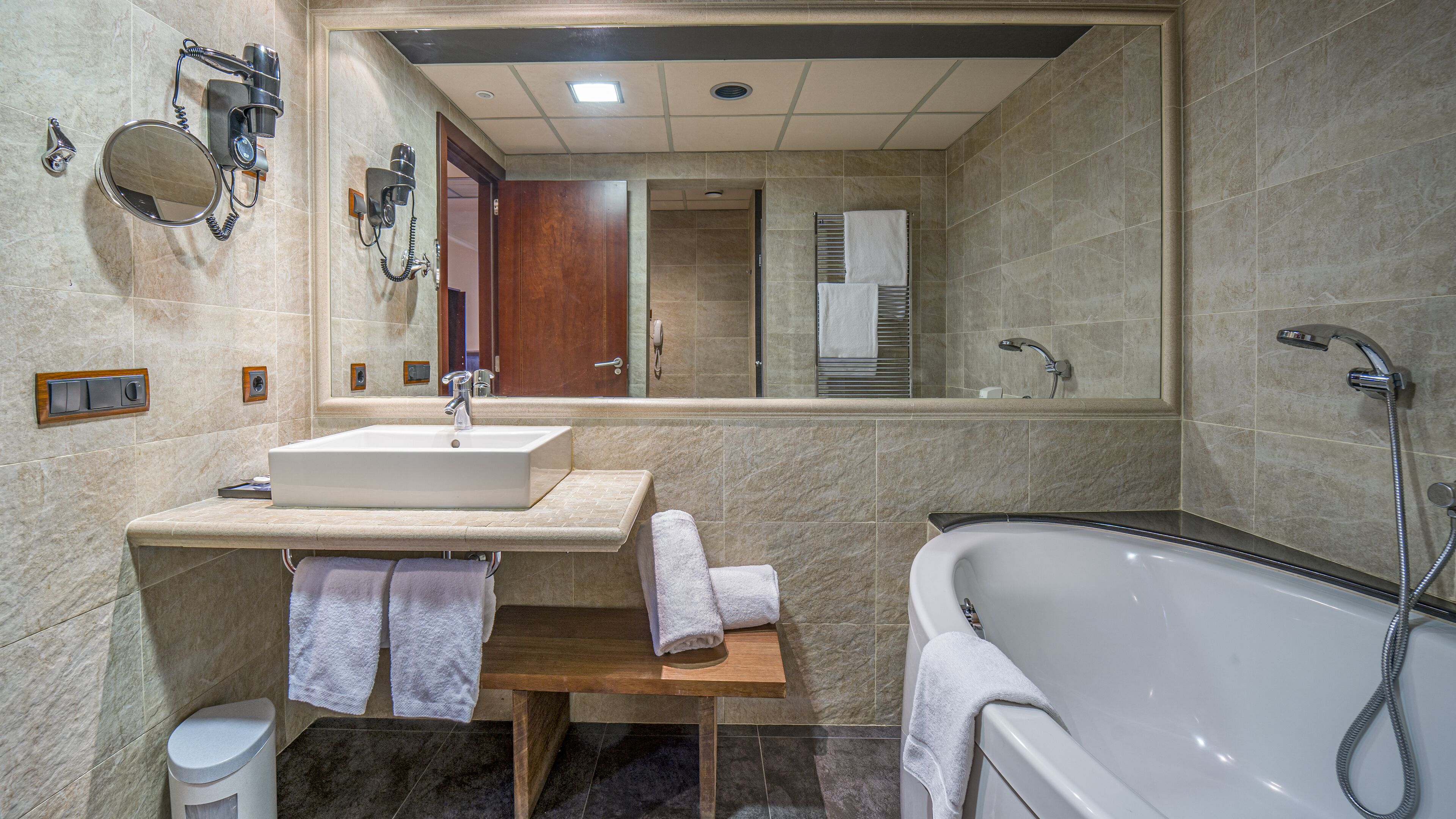 Une salle de bain moderne avec un grand miroir, un lavabo, un meuble-vasque, un porte-serviettes, un petit tabouret en bois, des toilettes et une baignoire équipée d’une douchette.