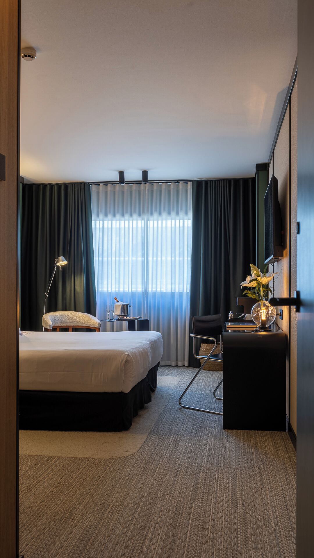 A hotel room with a bed, bedside table, lamp, chair, desk, and a large mirror.