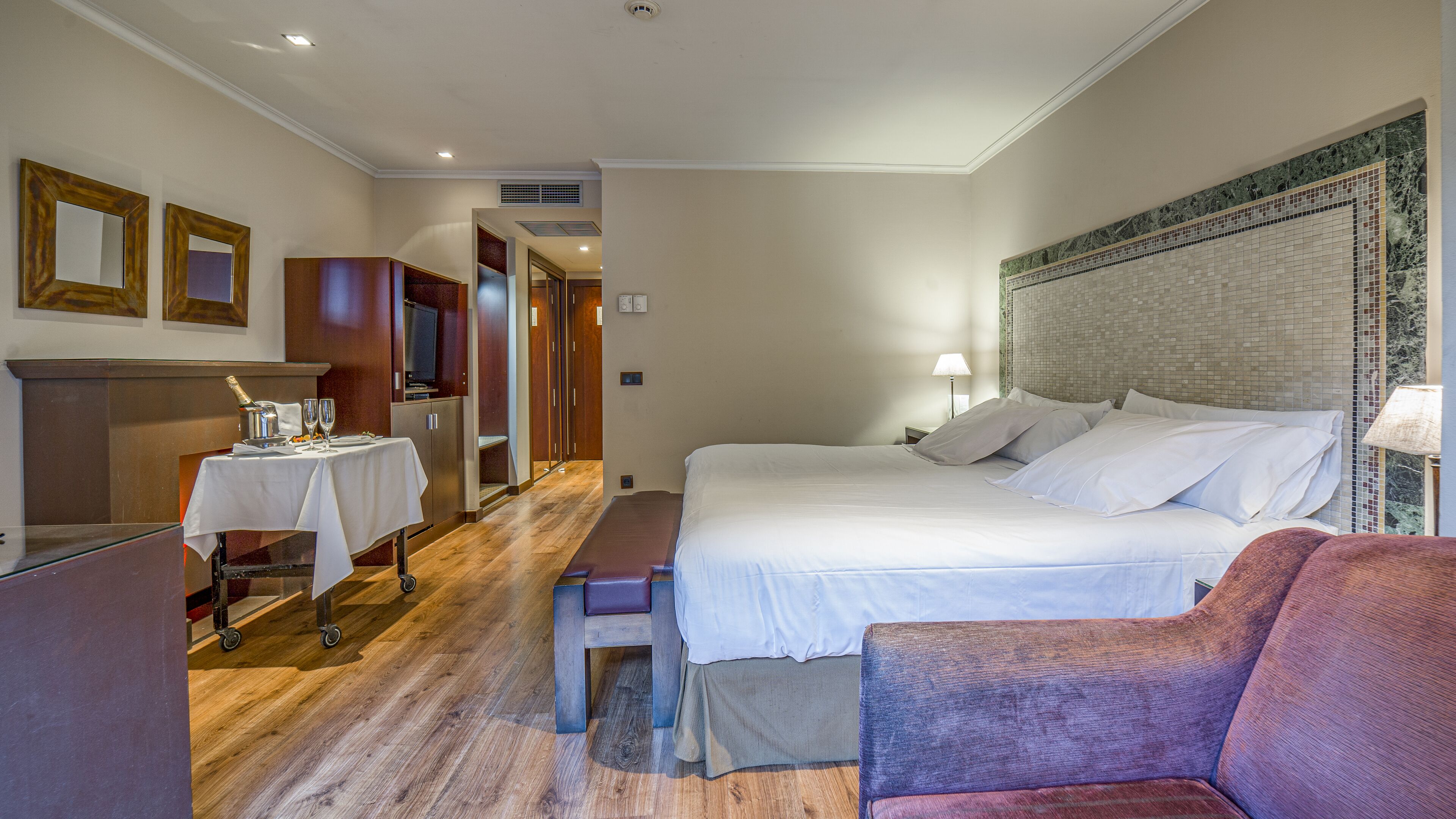 Habitación de hotel con cama, una mesita con botella y vasos, televisión y una zona de estar con un sillón.