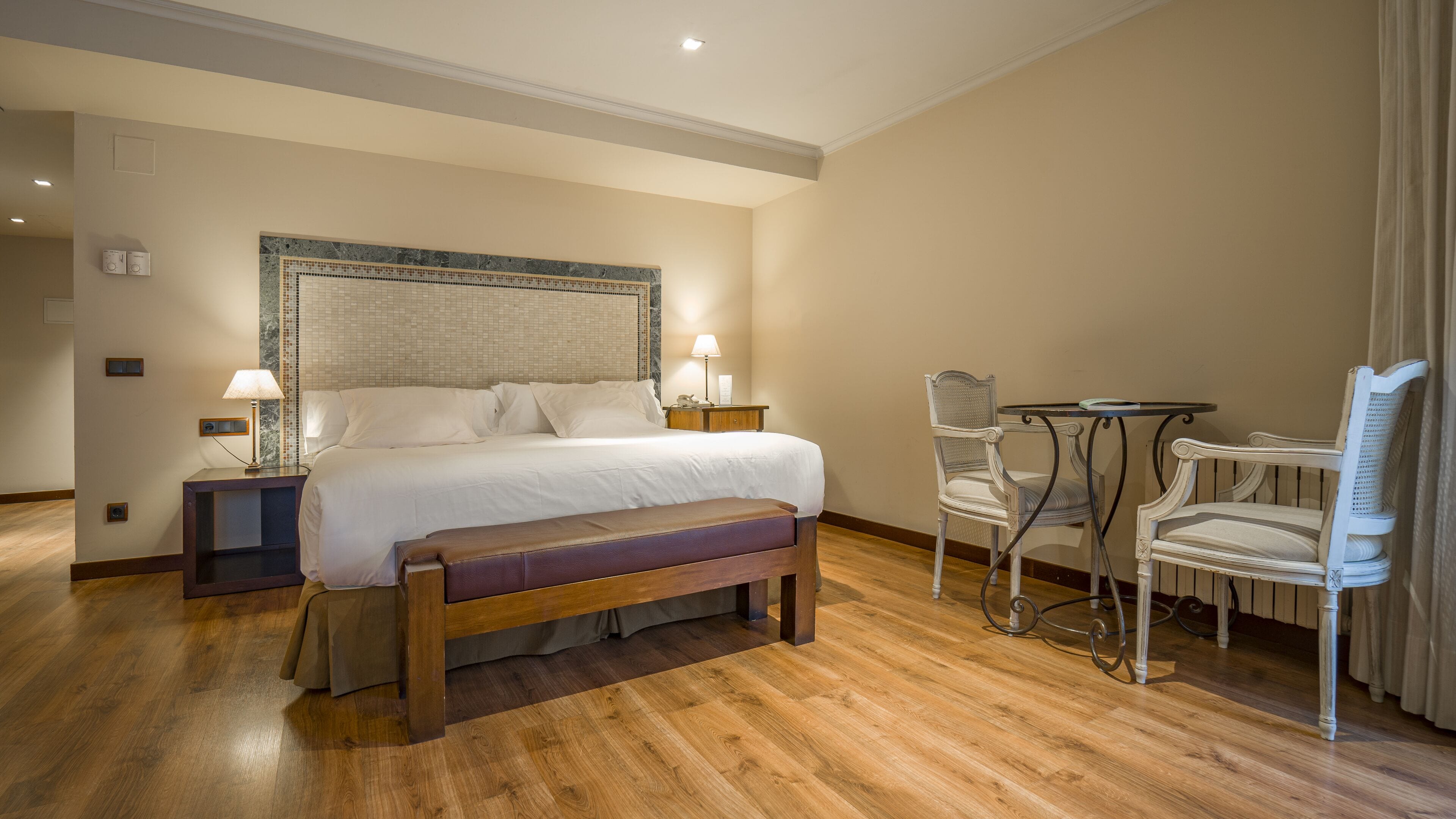A hotel room with a large bed, bedside tables with lamps, a wooden bench, a small round table with chairs, and a decorative wall panel.