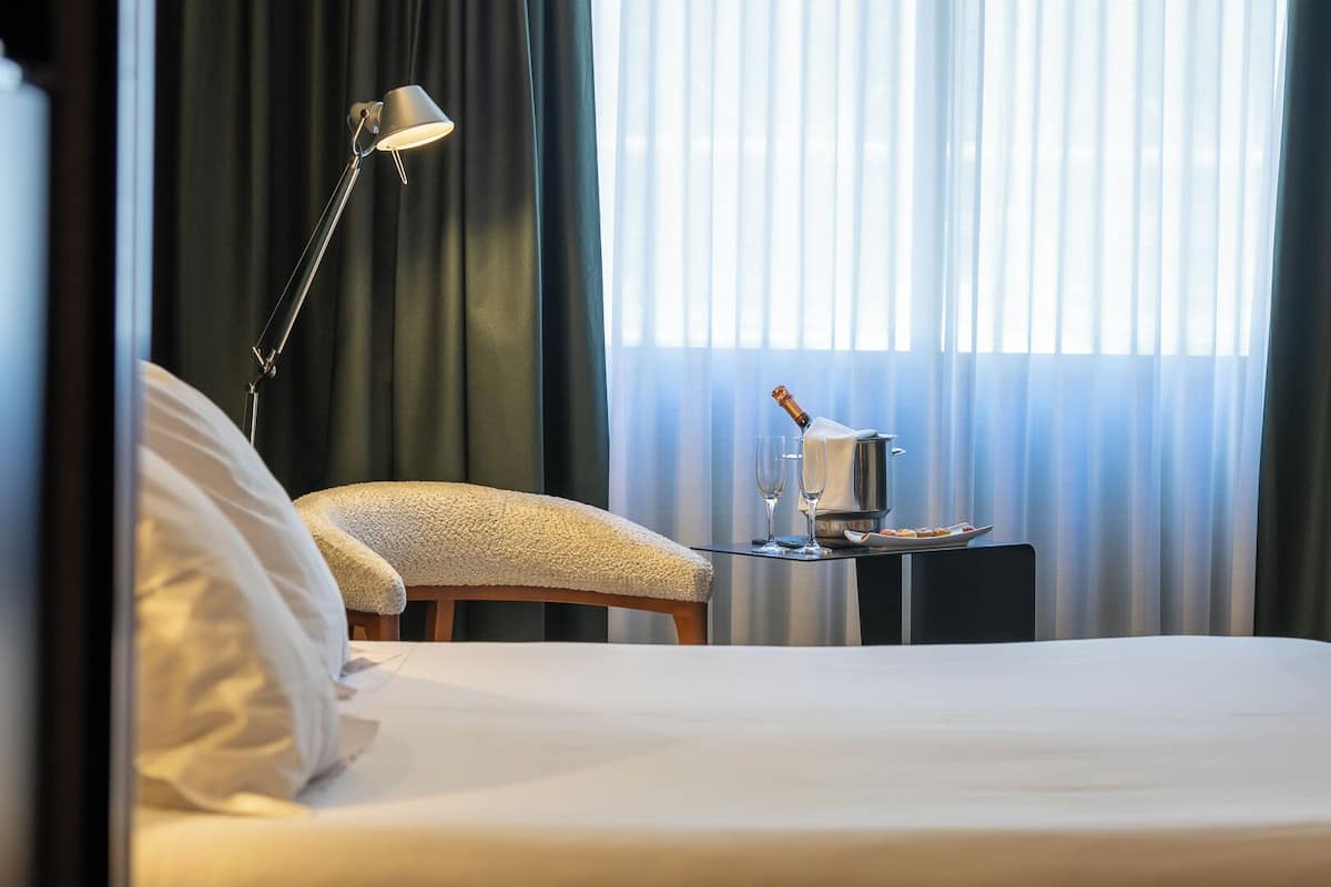 Quarto de hotel com cama, abajur, uma garrafa de champanhe e uma mesinha com um copo.