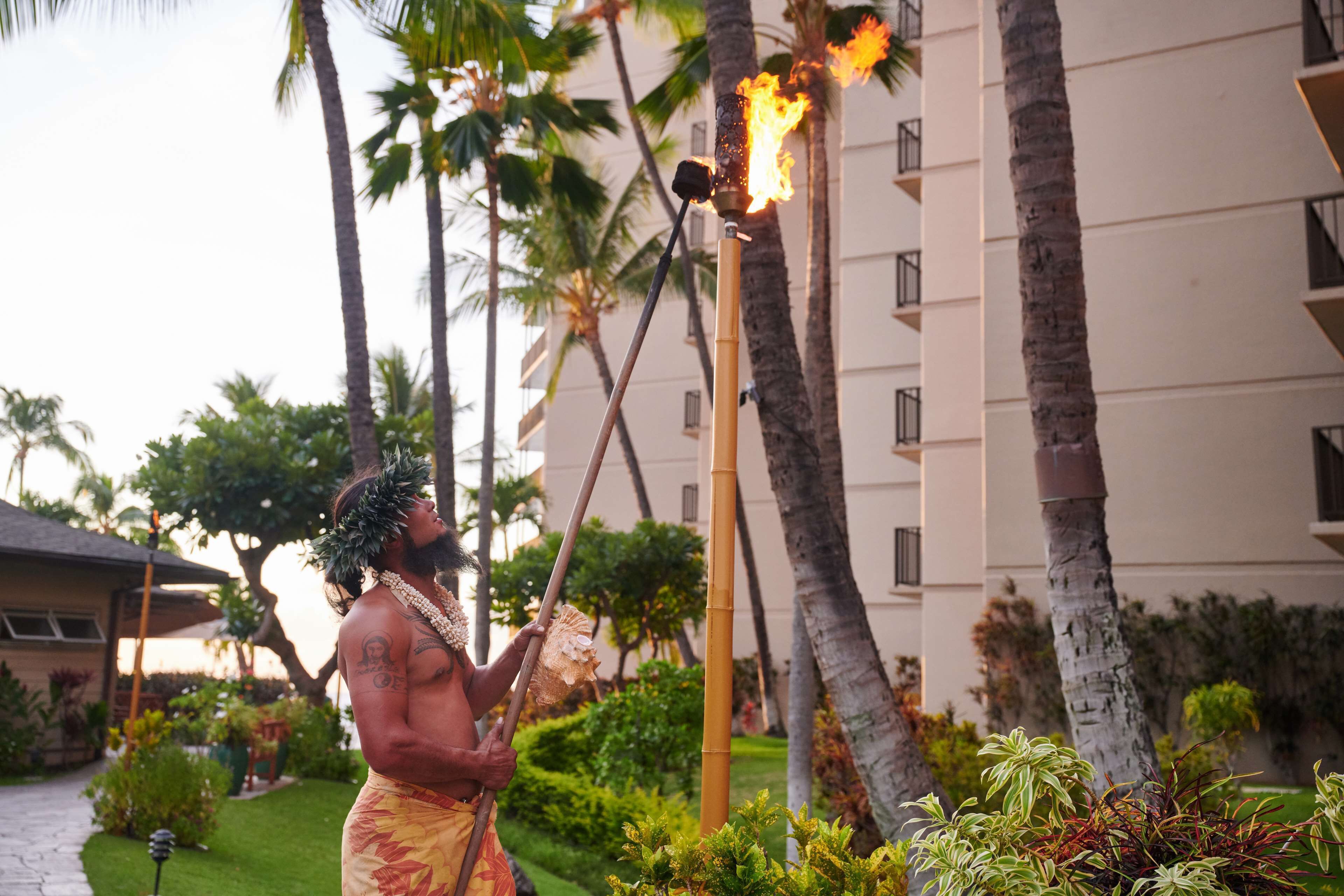 A person performing a torch ceremony in a tropical setting with palm trees and a building in the background.