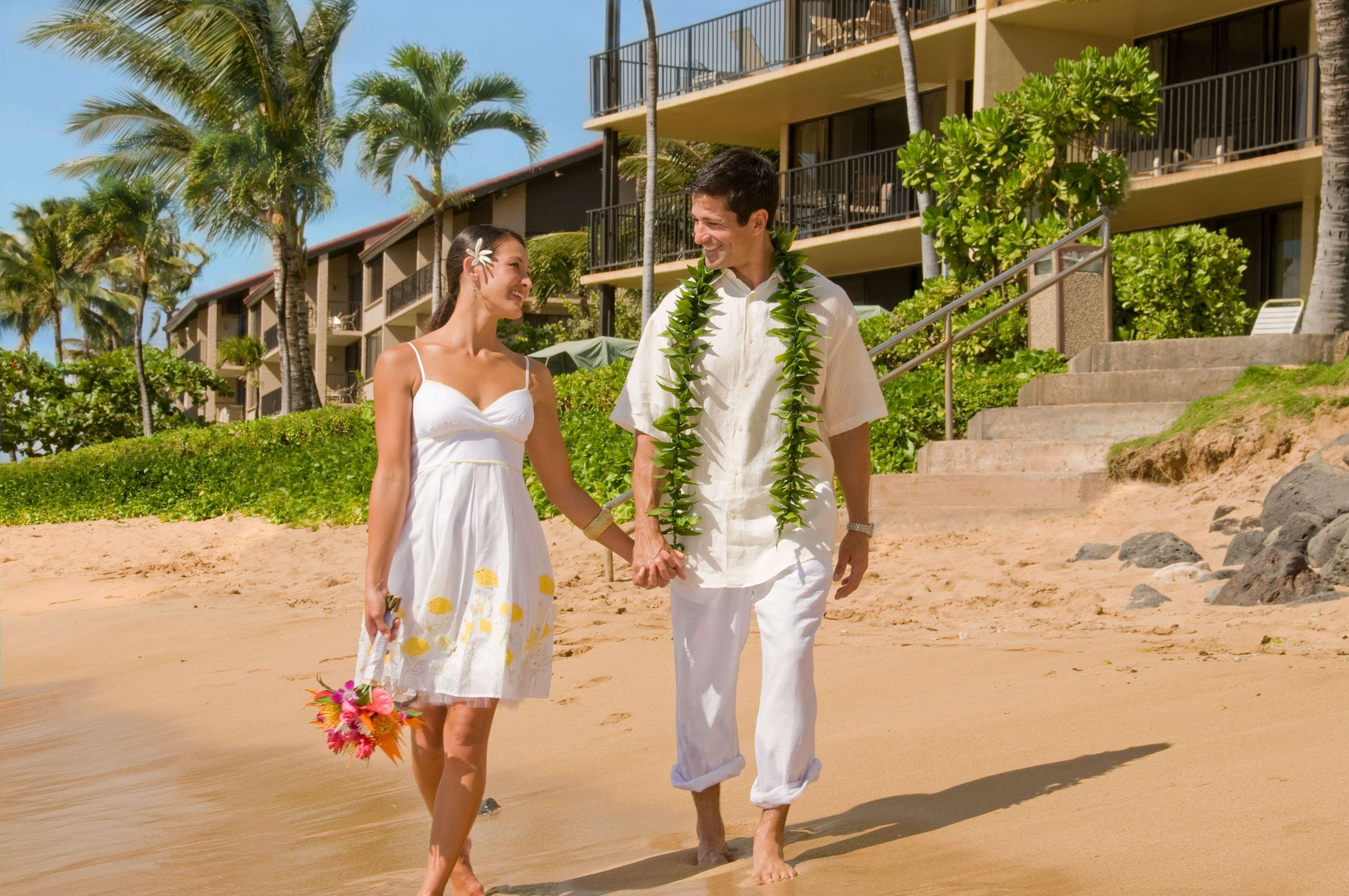 A couple holding hands on a sandy beach with a hotel in the background.