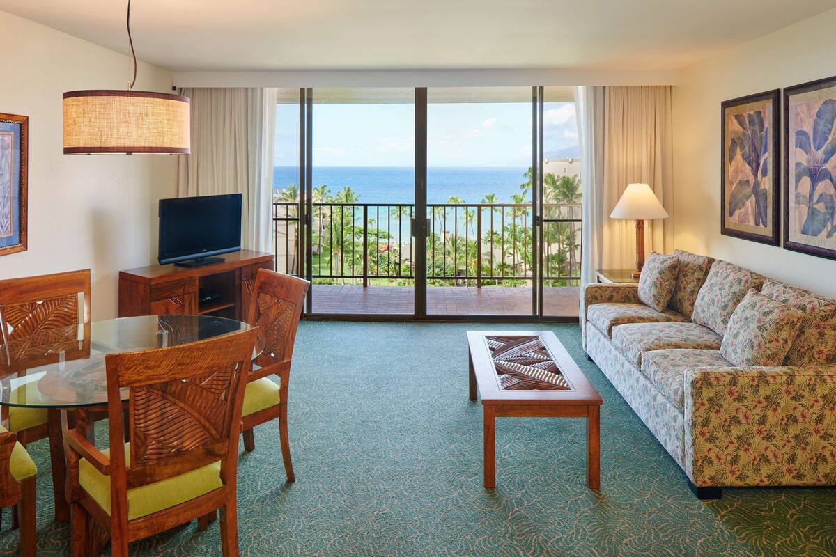 A hotel room with a balcony, a television, a sofa, and a dining area with chairs and a table.