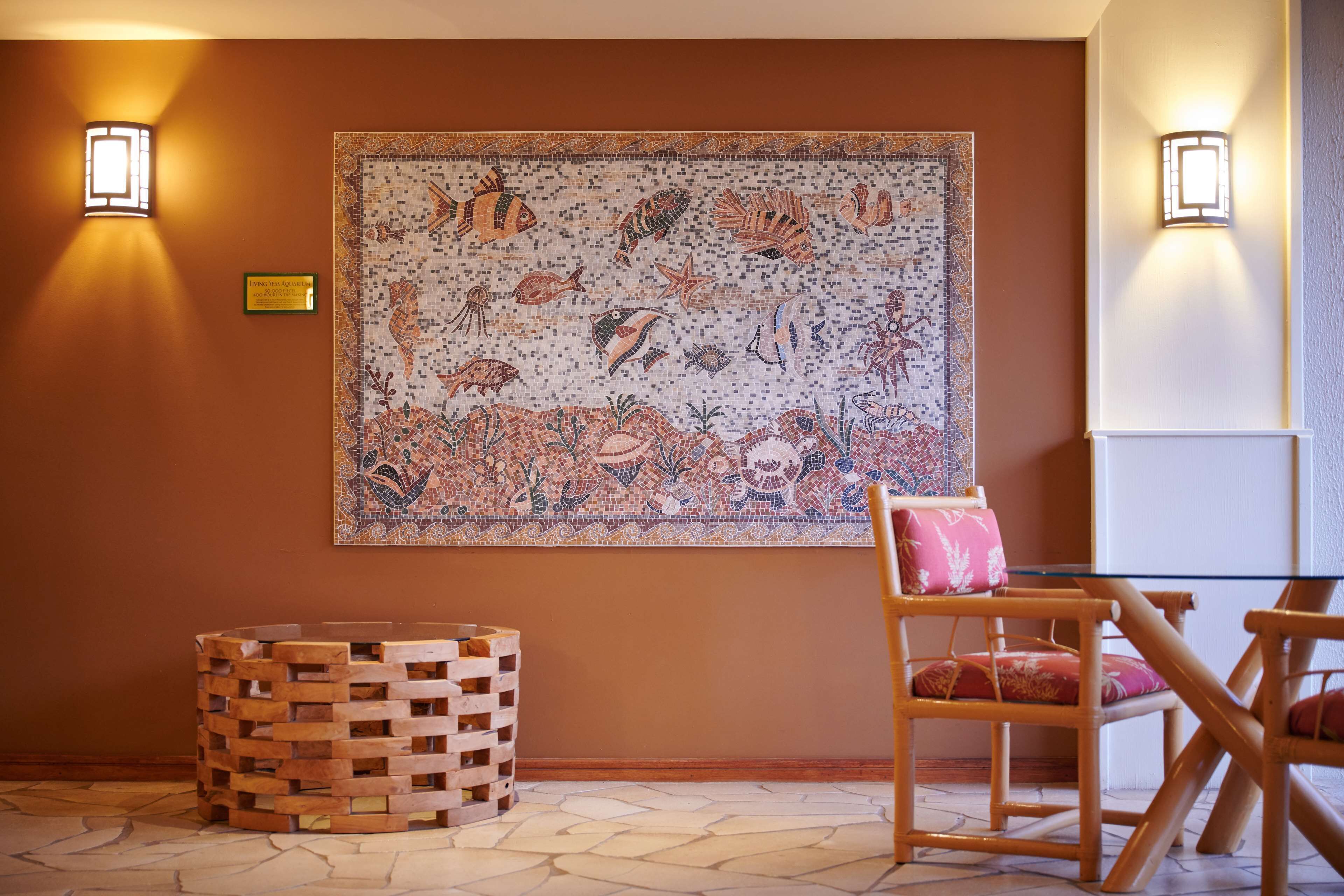 A room with a mosaic wall art, a wooden side table, and a wicker chair.