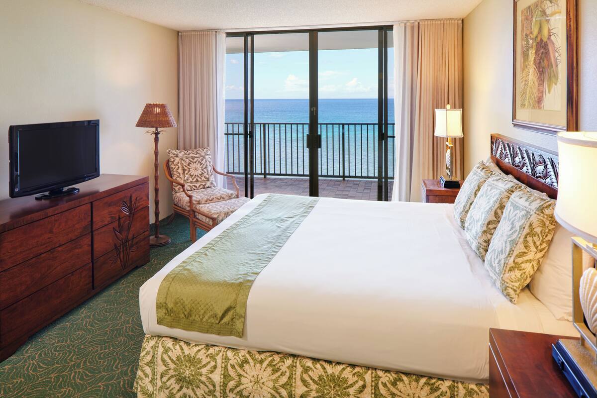 A hotel room with a large bed, a television on a wooden dresser, a chair, a balcony with a view of the ocean, and a painting on the wall.