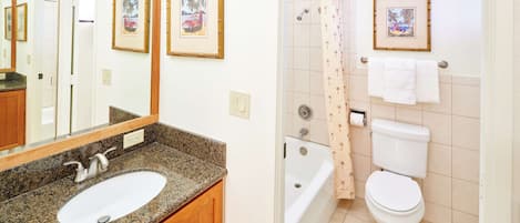 A bathroom with a granite countertop sink, wooden cabinets, a white toilet, and a shower with a bamboo shower curtain.