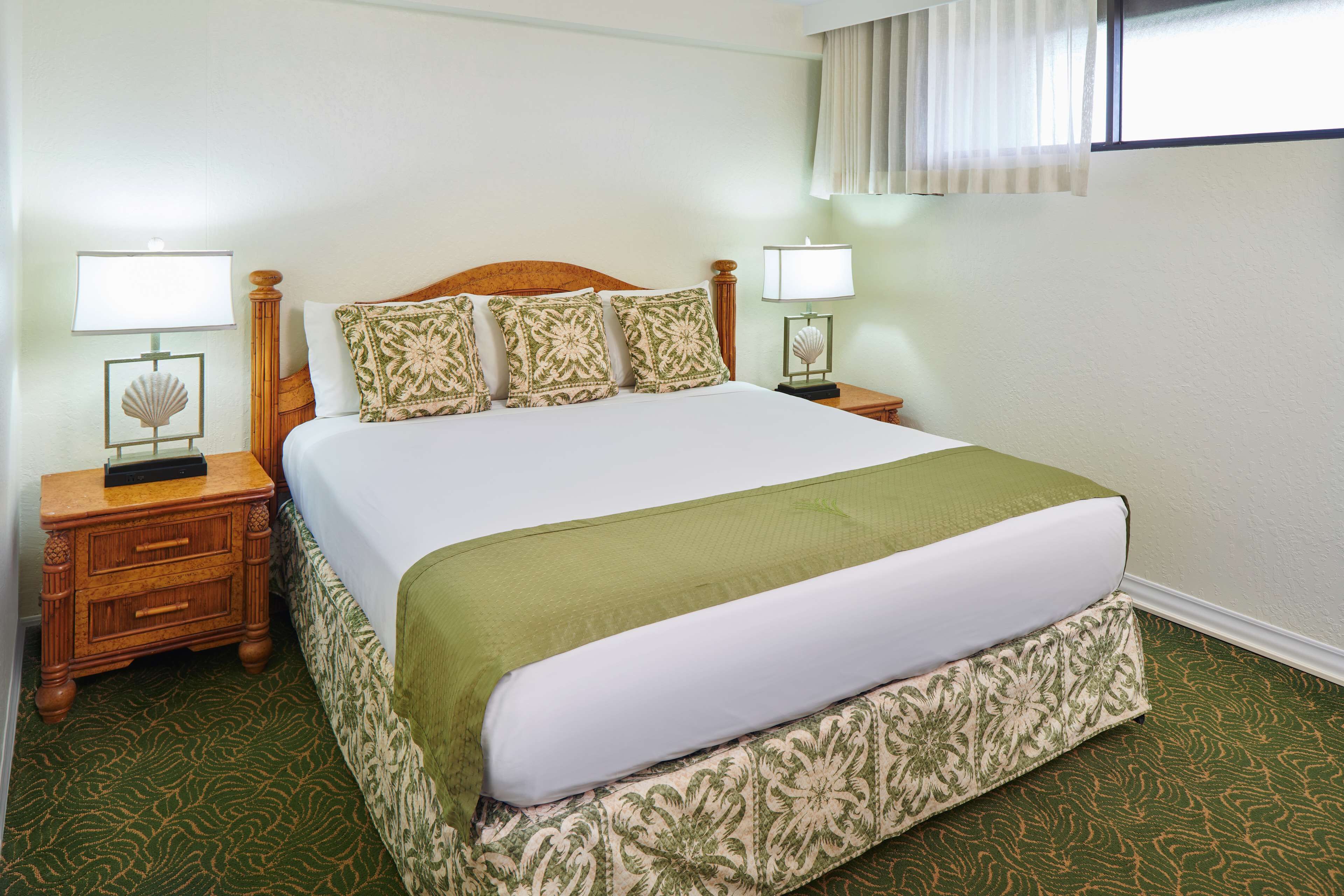 A neatly made bed with a green bedspread and matching pillows, two bedside tables with lamps, and a patterned carpet.