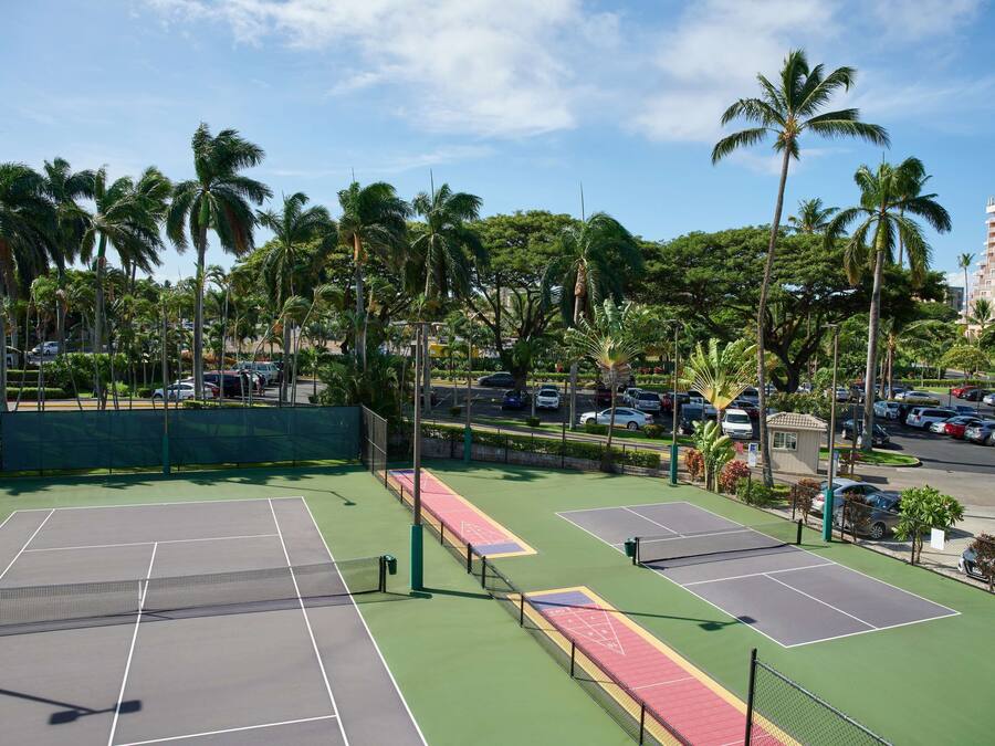 Un court de tennis comprenant plusieurs terrains, entouré de palmiers et doté d’un espace de stationnement.