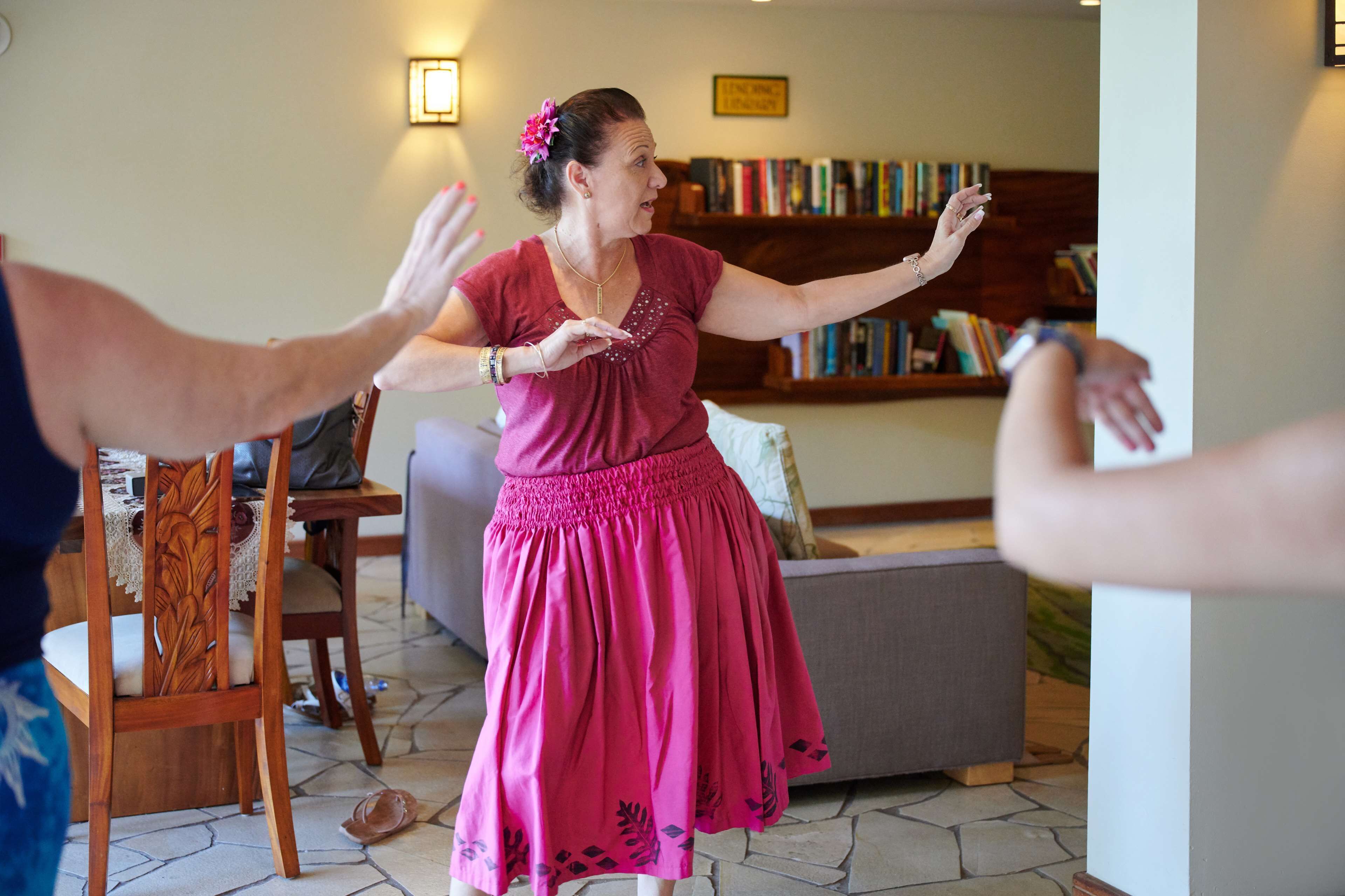 Una persona amb un vestit rosa ballant en un espai amb una cadira de fusta, un sofà i una prestatgeria al fons.
