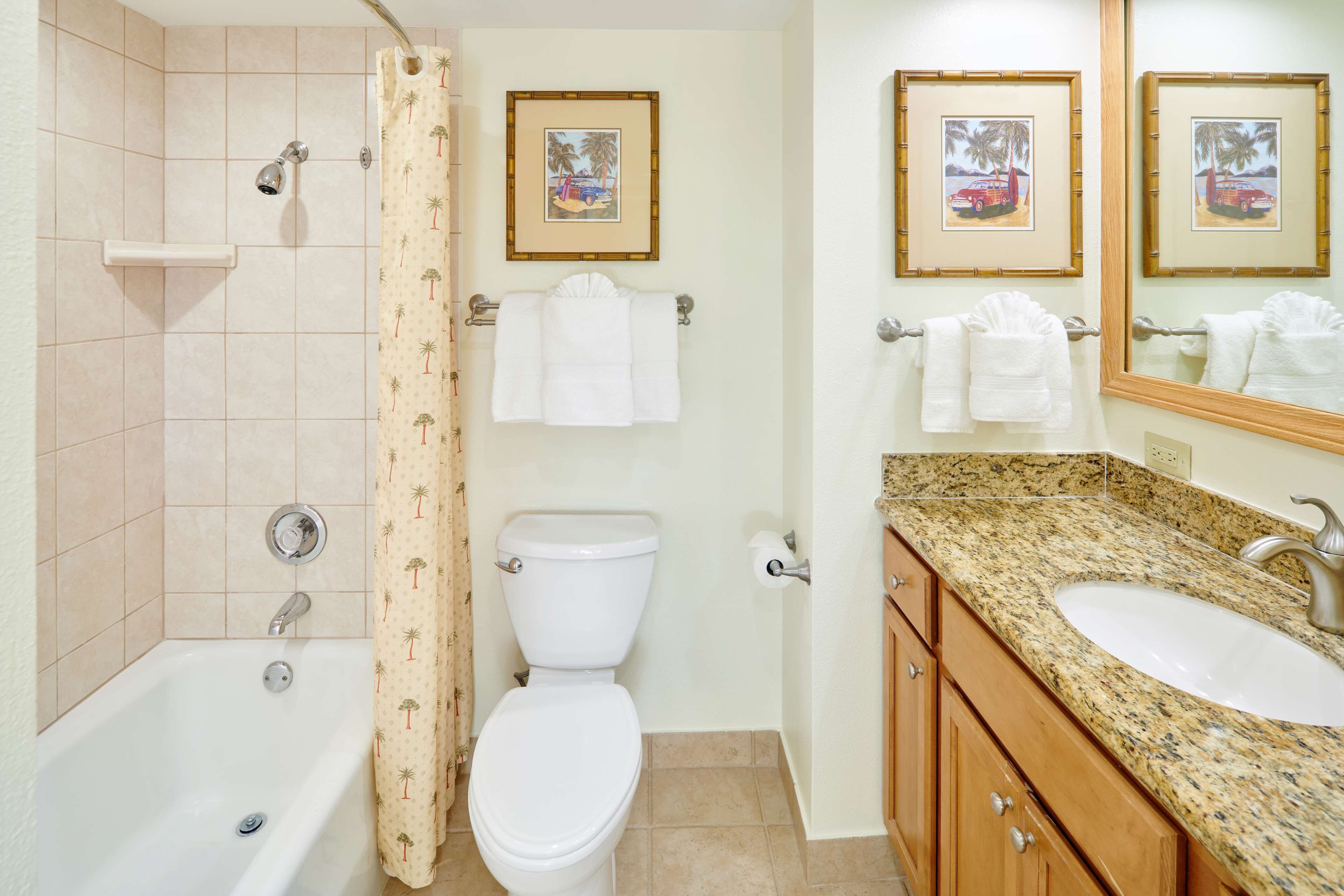 A bathroom with a bathtub, toilet, and sink, featuring framed pictures on the wall and a shower curtain with palm tree patterns.